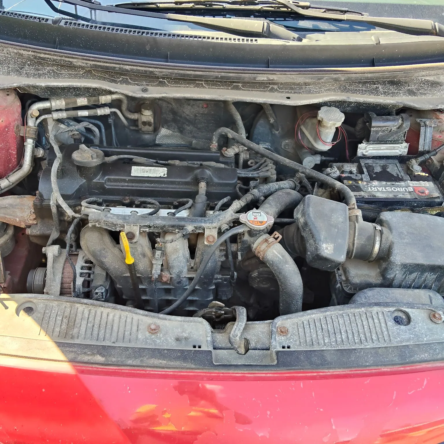 Widok z góry na komorę silnika w czerwonym samochodzie. Widoczny jest zakurzony silnik spalinowy z różnymi przewodami, rurami i elementami mechanicznymi. Po prawej stronie znajduje się czarny akumulator z napisem 'EUROSTART'. W dolnej części zdjęcia widoczny jest fragment czerwonego zderzaka.