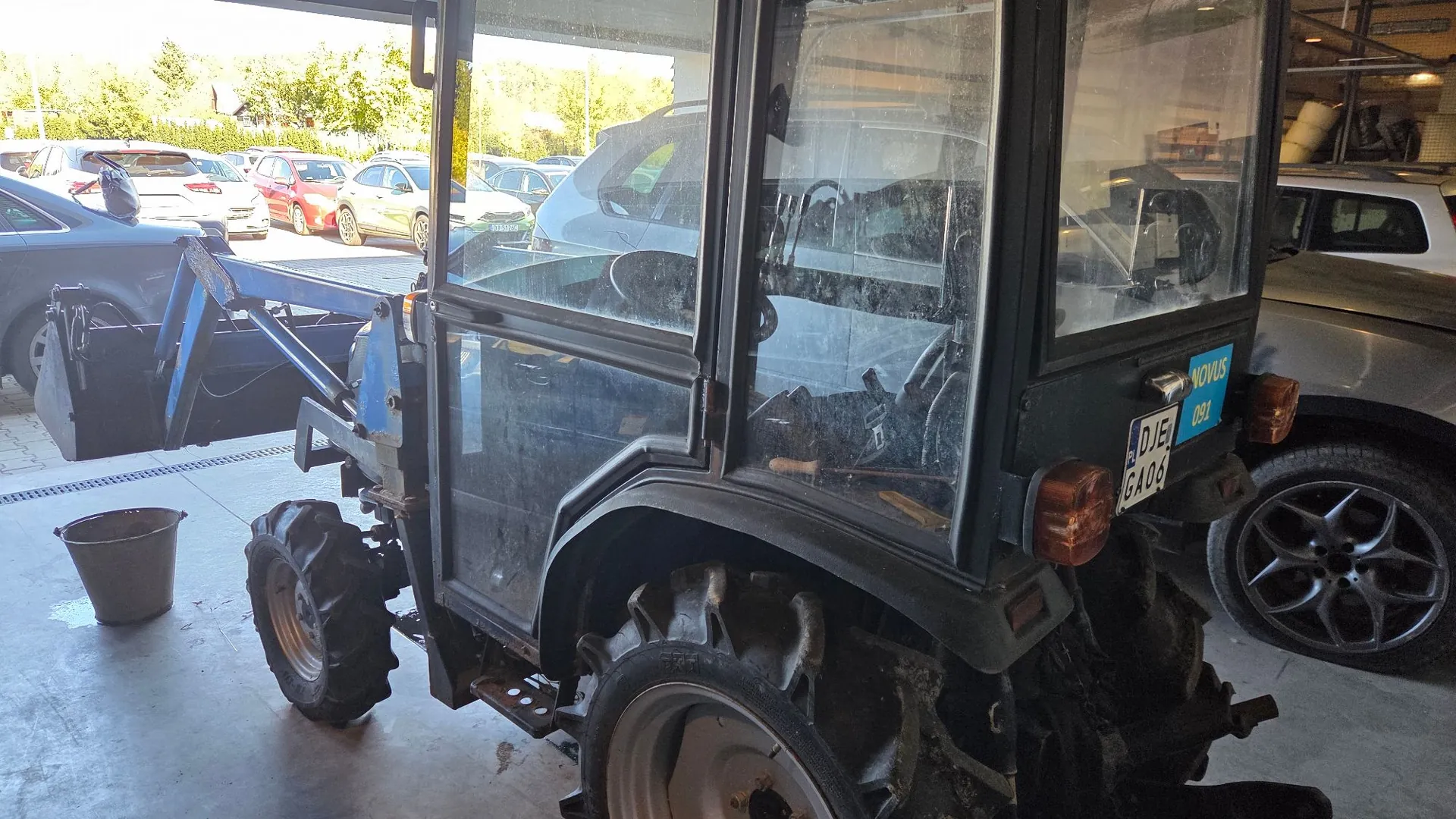 Zdjęcie przedstawia niebieski traktor z przeszkloną kabiną, zaparkowany w garażu lub hali. Traktor jest widoczny z boku i z tyłu. W tle, za traktorem, widoczne są zaparkowane samochody osobowe w różnych kolorach: srebrny, czerwony, żółty i szary. Po prawej stronie znajduje się fragment ciemnego samochodu terenowego. Na tylnej części traktora widoczna jest niebieska tabliczka z napisem 'NOVUS 081' oraz tablica rejestracyjna 'DJE GA04'. Na podłodze obok traktora stoi szary plastikowy kubeł.