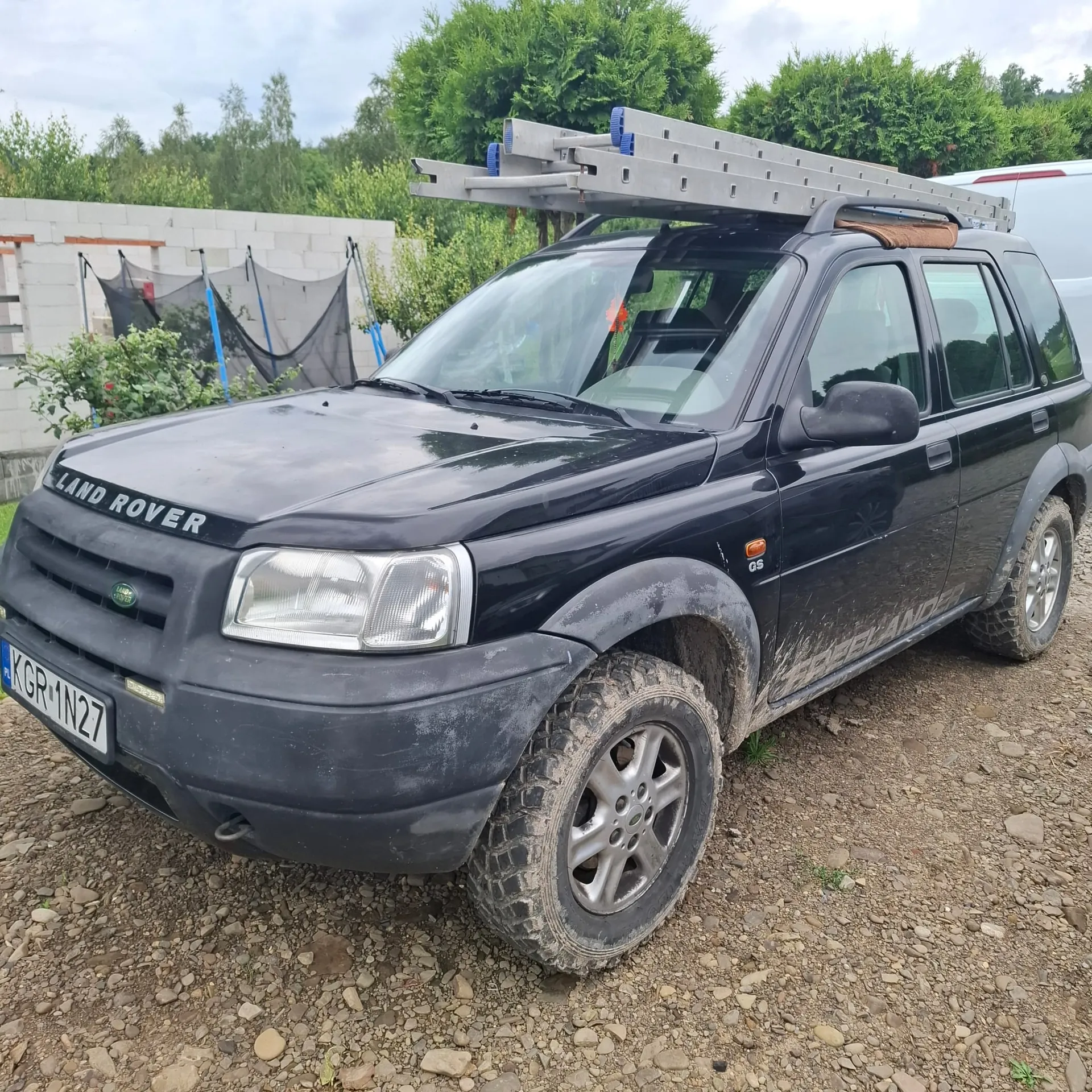 Czarny samochód terenowy marki Land Rover z zamontowaną na dachu drabiną aluminiową. Pojazd jest zabrudzony błotem, szczególnie w dolnych partiach nadwozia i na oponach. Samochód stoi na żwirowym podłożu. Widoczna jest tablica rejestracyjna o numerze KGR 1N27 oraz emblemat GS na przednim błotniku.