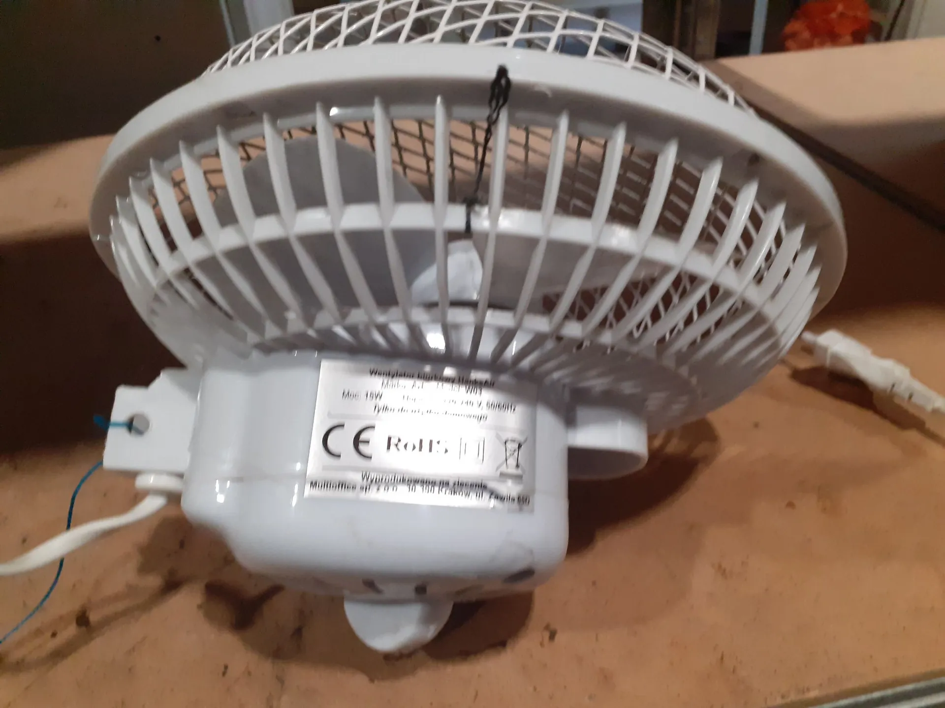 Biały wentylator łazienkowy z plastikową osłoną kratką. Na obudowie silnika znajduje się etykieta z tekstem: 'Wentylator łazienkowy', 'Moc: 18W', '230V 50/60Hz', oraz oznaczeniami CE i RoHS. Z urządzenia wychodzą niebieski i biały przewody elektryczne.