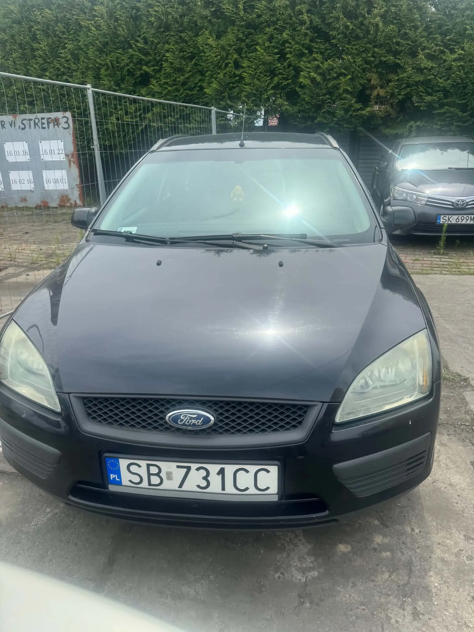 Czarny samochód marki Ford widziany od przodu, zaparkowany na betonowym podłożu. Pojazd posiada polską tablicę rejestracyjną o numerze SB 731CC. W tle widoczny jest metalowy płot z napisem 'STREFA 3' oraz zielony żywopłot. Po prawej stronie częściowo widoczny jest inny, srebrny samochód z tablicą SK 699M.
