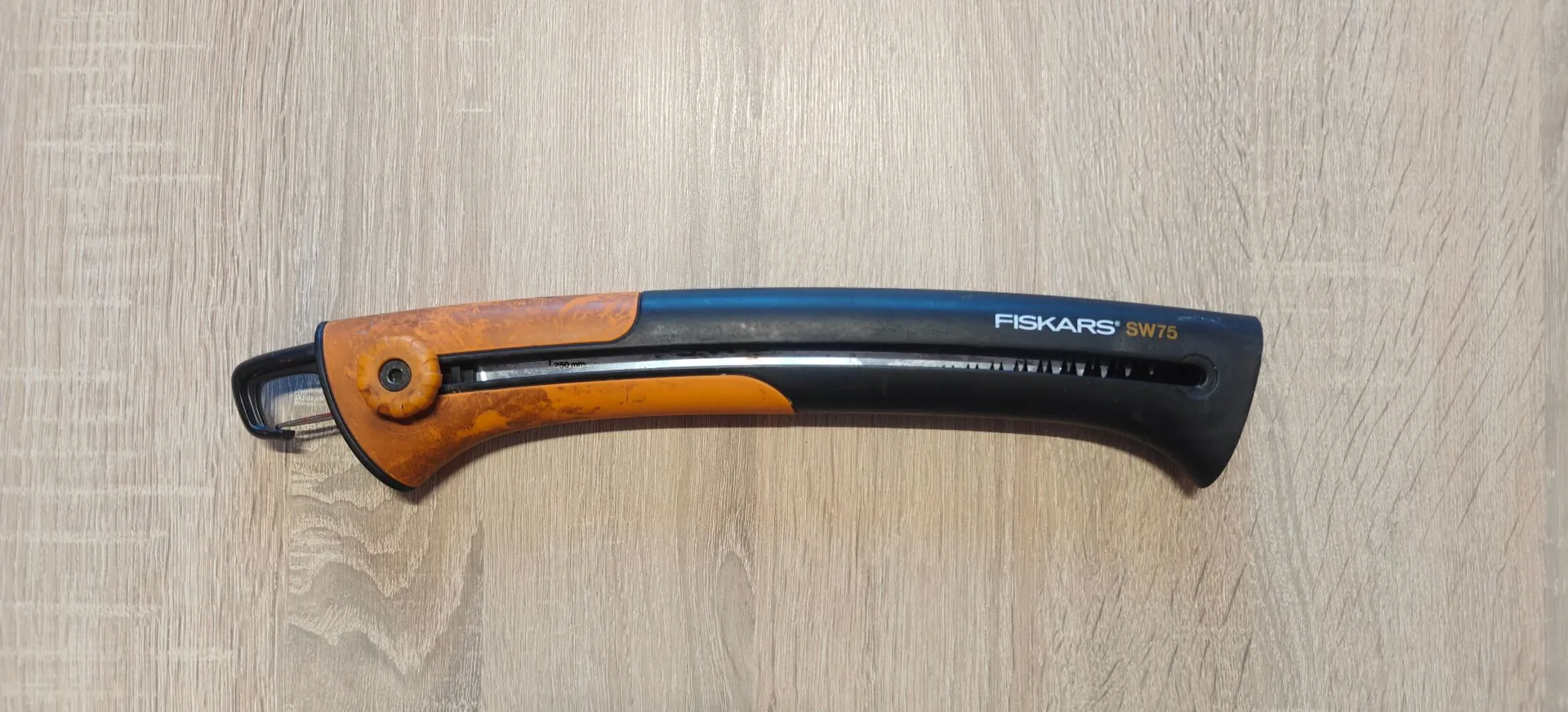 Piła ręczna marki Fiskars model SW75 leżąca poziomo na jasnym, drewnianym blacie. Narzędzie posiada czarną rękojeść z napisem 'FISKARS SW75' oraz pomarańczowe elementy wykończenia. Ostrze piły jest częściowo schowane w obudowie.