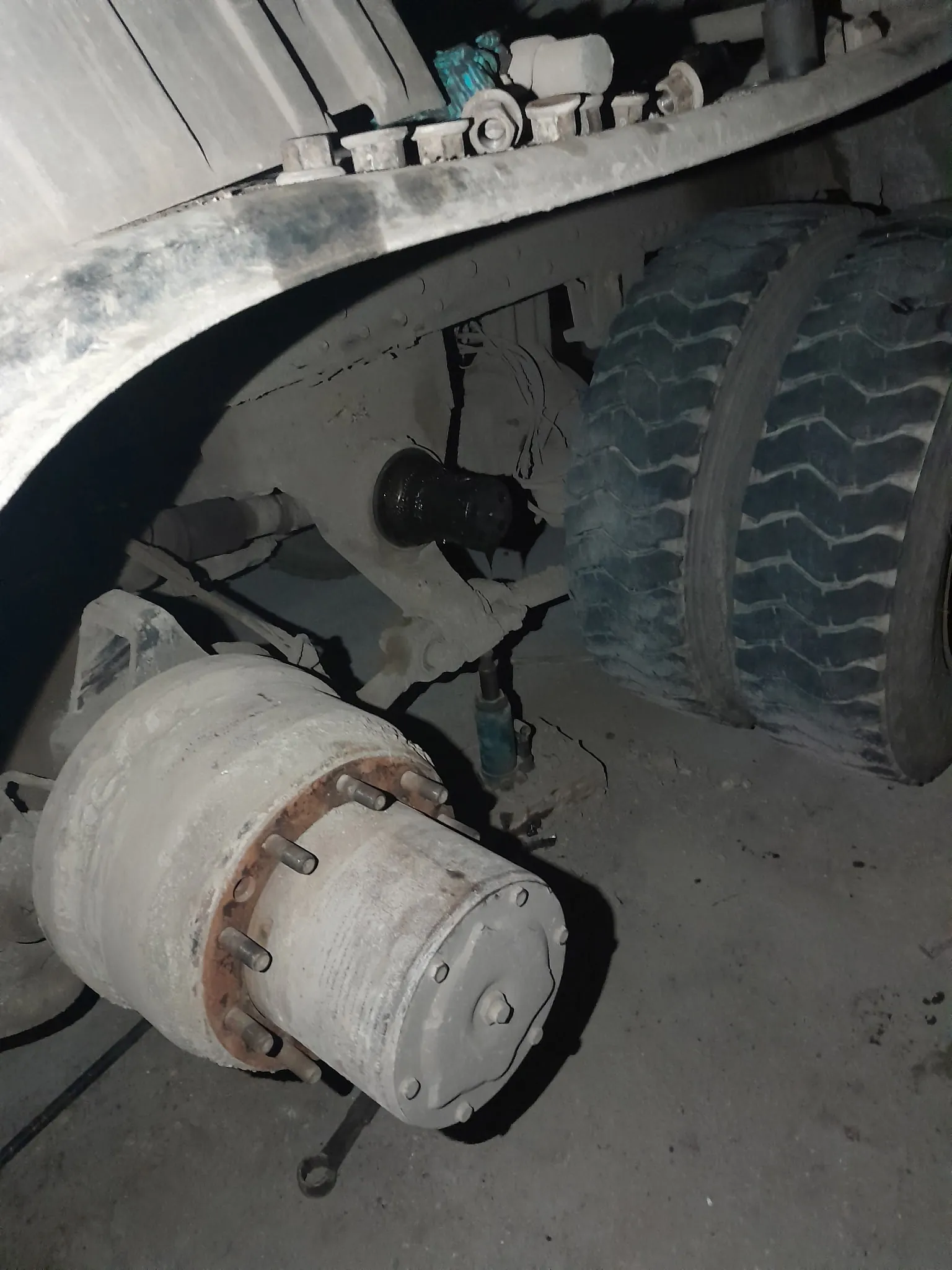 Zdjęcie przedstawia fragment podwozia pojazdu ciężarowego. W centrum widoczny jest duży, cylindryczny element mechaniczny z mocowanymi śrubami i śladami rdzy na brzegu. Po prawej stronie znajdują się dwie duże opony z głębokim, ząbkowanym profilem. Na górze obrazu widoczna jest metalowa rama pojazdu z kilkoma małymi elementami mechanicznymi, w tym niebieskim komponentem. Na betonowej podłodze leży klucz. Całość pokryta jest kurzem i śladami zużycia.