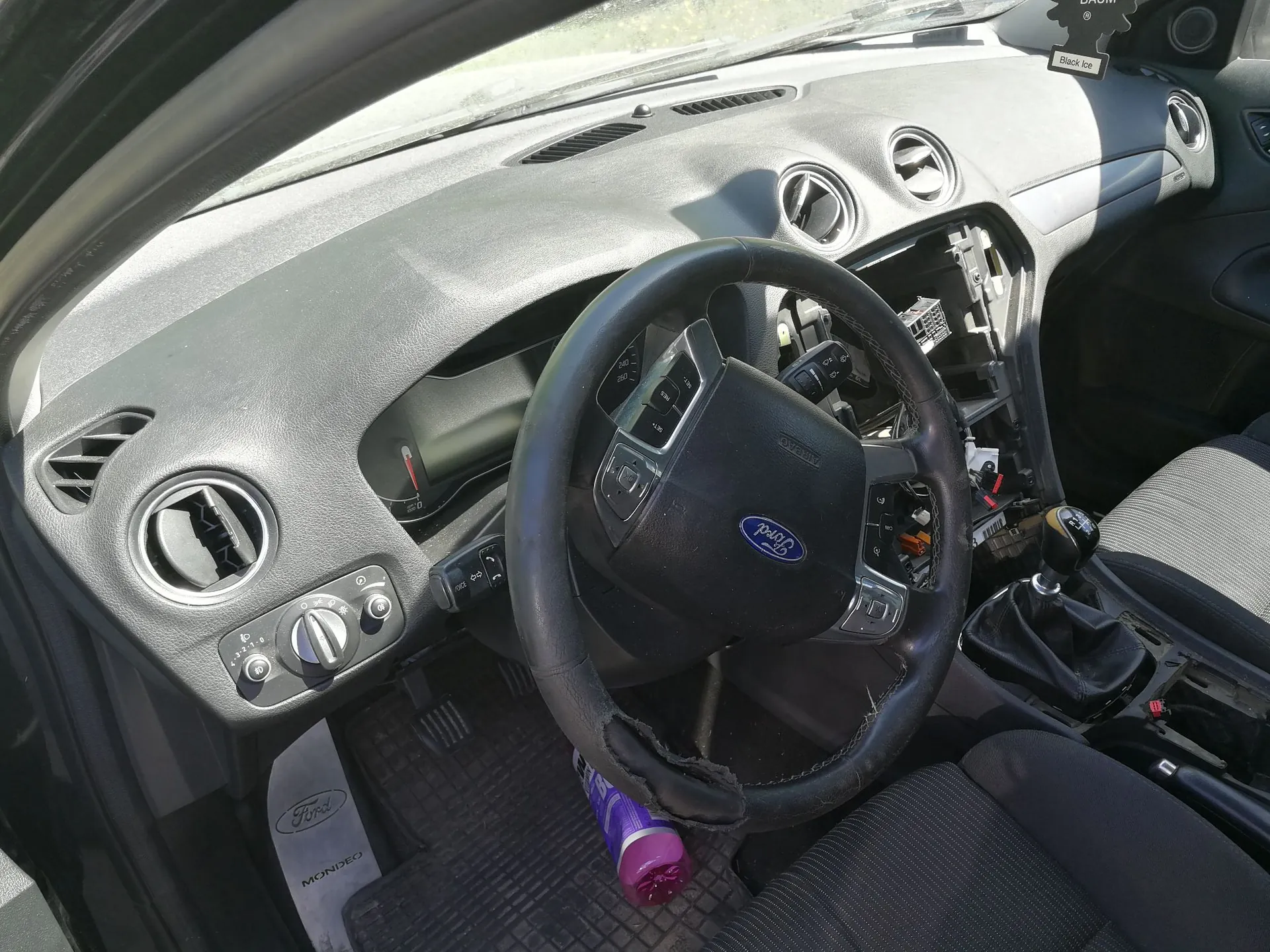Wnętrze samochodu marki Ford, widok z perspektywy kierowcy. Widoczna jest czarna deska rozdzielcza, kierownica z logo Ford oraz fotele. W centralnej części deski rozdzielczej brakuje radia/panelu sterowania, odsłaniając okablowanie i wnętrze schowka. Na podłodze po stronie kierowcy leży fioletowa butelka oraz mata z napisem 'Ford MONDEO'.