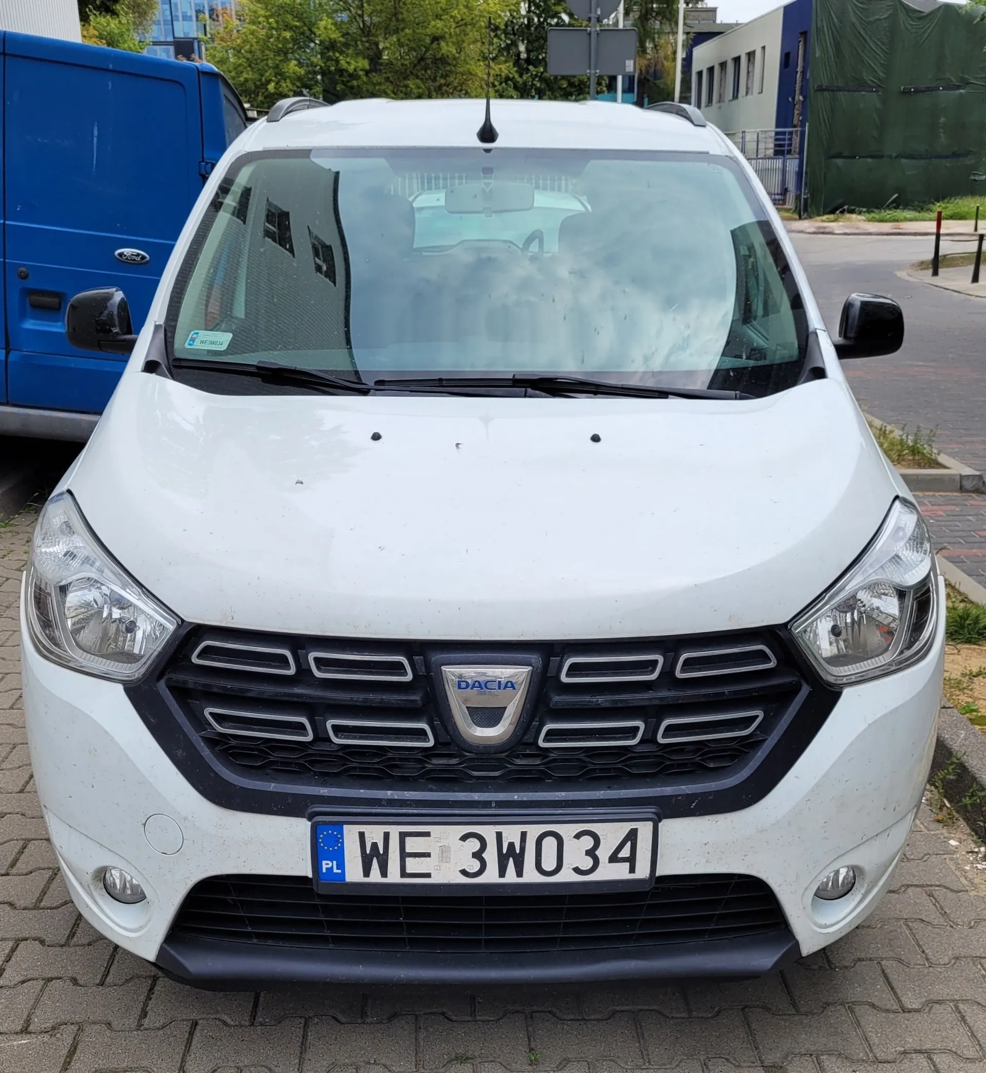 Widok z przodu białego samochodu marki Dacia zaparkowanego na kostce brukowej. Pojazd posiada polską tablicę rejestracyjną o numerze WE 3WO34. W tle widoczny jest niebieski furgon oraz budynki.