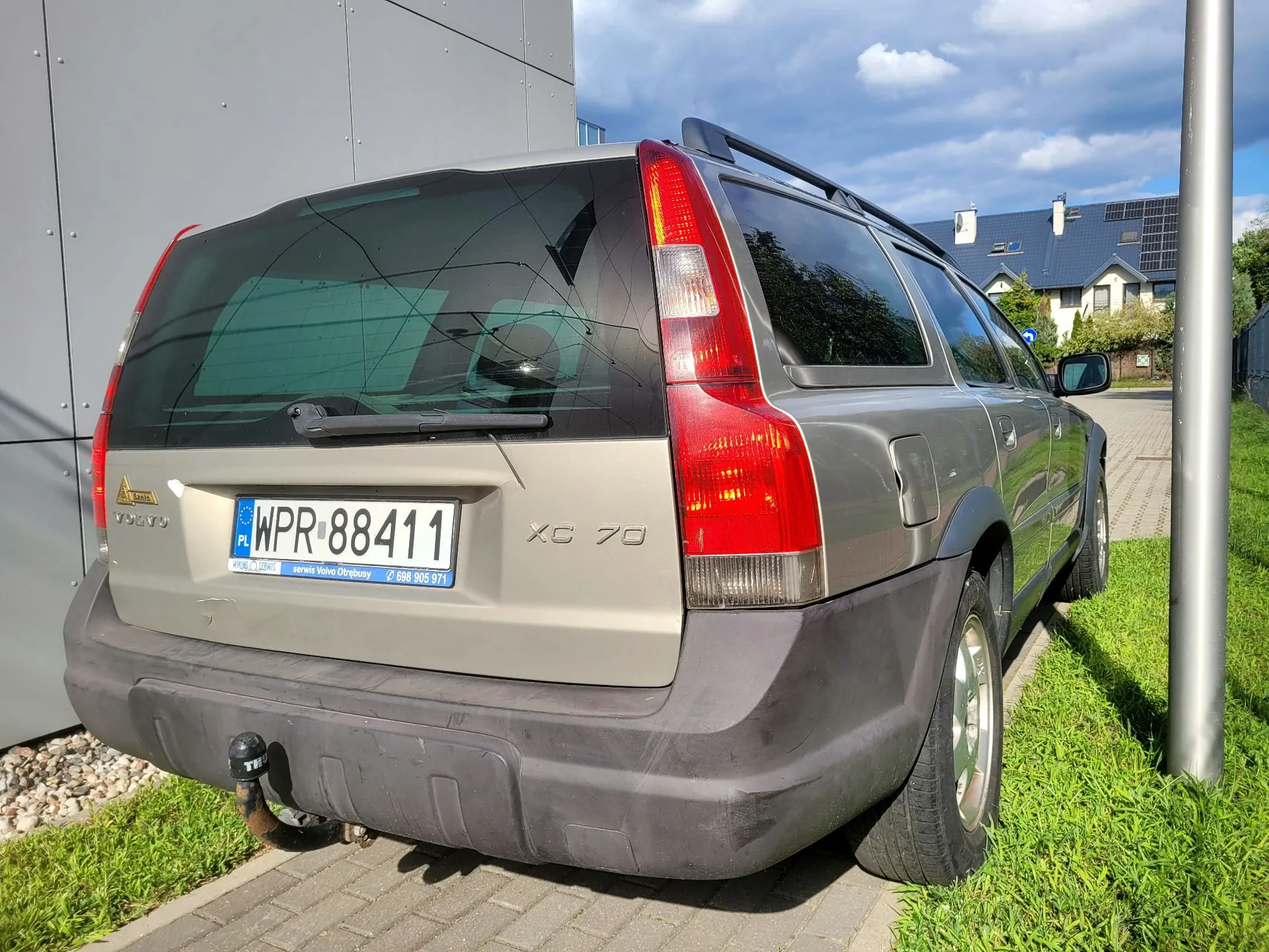 Srebrny samochód marki Volvo XC70 zaparkowany na kostce brukowej obok trawnika i szarego budynku. Widoczny jest tył pojazdu z tablicą rejestracyjną WPR 88411, napisem XC 70 oraz hakiem holowniczym. W tle widać domy jednorodzinne i niebo z chmurami.