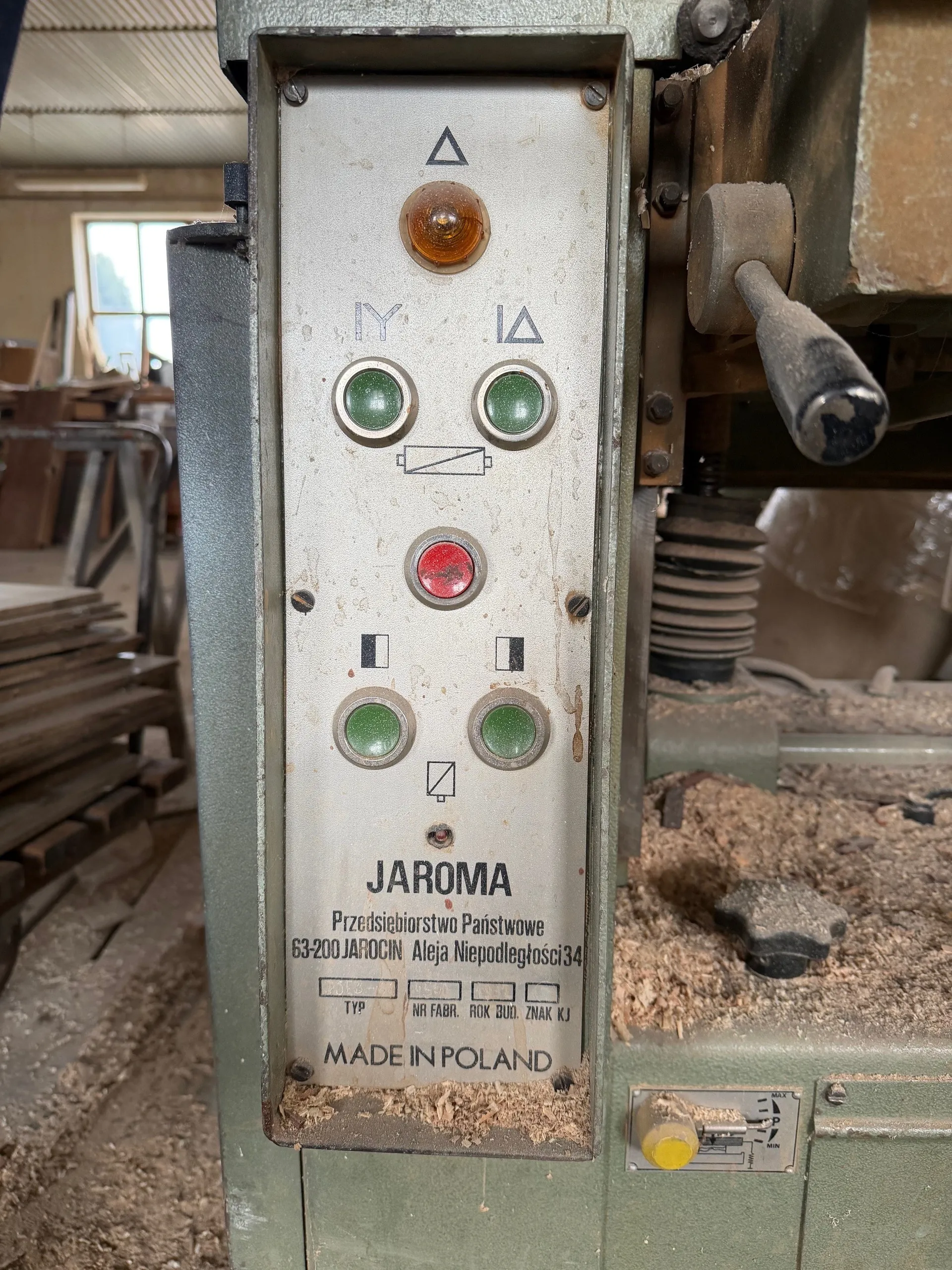 Zbliżenie na metalową deskę sterowniczą maszyny w warsztacie. Deska zawiera żółte światło ostrzegawcze, cztery zielone przyciski, czerwony przycisk oraz symbole: trójkąt, litery IY i IA, prostokąt z przekreślonym pionowym segmentem. Poniżej widoczny tekst: JAROMA, Przedsiębiorstwo Państwowe, 63-200 JAROCIN Aleja Niepodległości 34, TYP, NR FABR., ROK BUD., ZNAK KJ oraz MADE IN POLAND. W tle warsztat z drewnianymi deskami i wiórami.