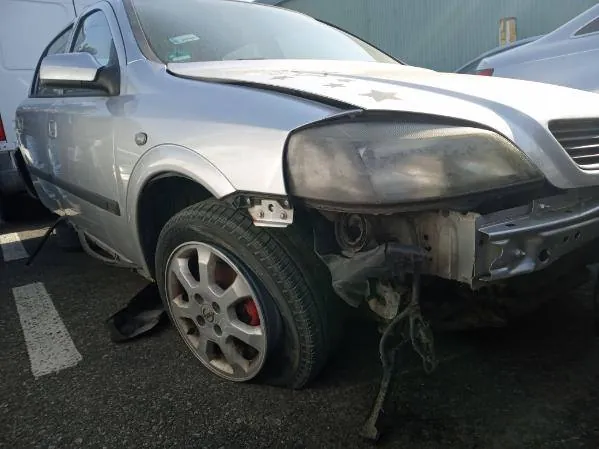 Zdjęcie przedstawia srebrny samochód osobowy z poważnymi uszkodzeniami przedniej części. Widoczne są zdeformowane maskę, zniszczony zderzak, uszkodzony reflektor oraz odsłonięte elementy podwozia. Koło przednie z aluminiową felgą znajduje się w pozycji naturalnej, a na asfalcie widoczna jest biała linia parkingu. W tle częściowo widoczny jest inny pojazd. Brak czytelnego tekstu na obrazie.