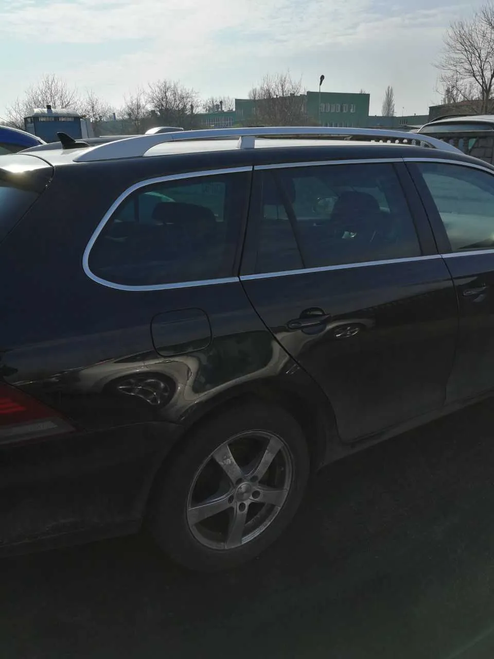 Czarny samochód typu kombi z białymi dachówkami dachowymi, przyciemnianymi szybami i srebrnymi felgami. Samochód zaparkowany na zewnątrz, na tle budynków, drzew bez liści i częściowo zachmurzonego nieba. Widoczne są również inne pojazdy w tle.