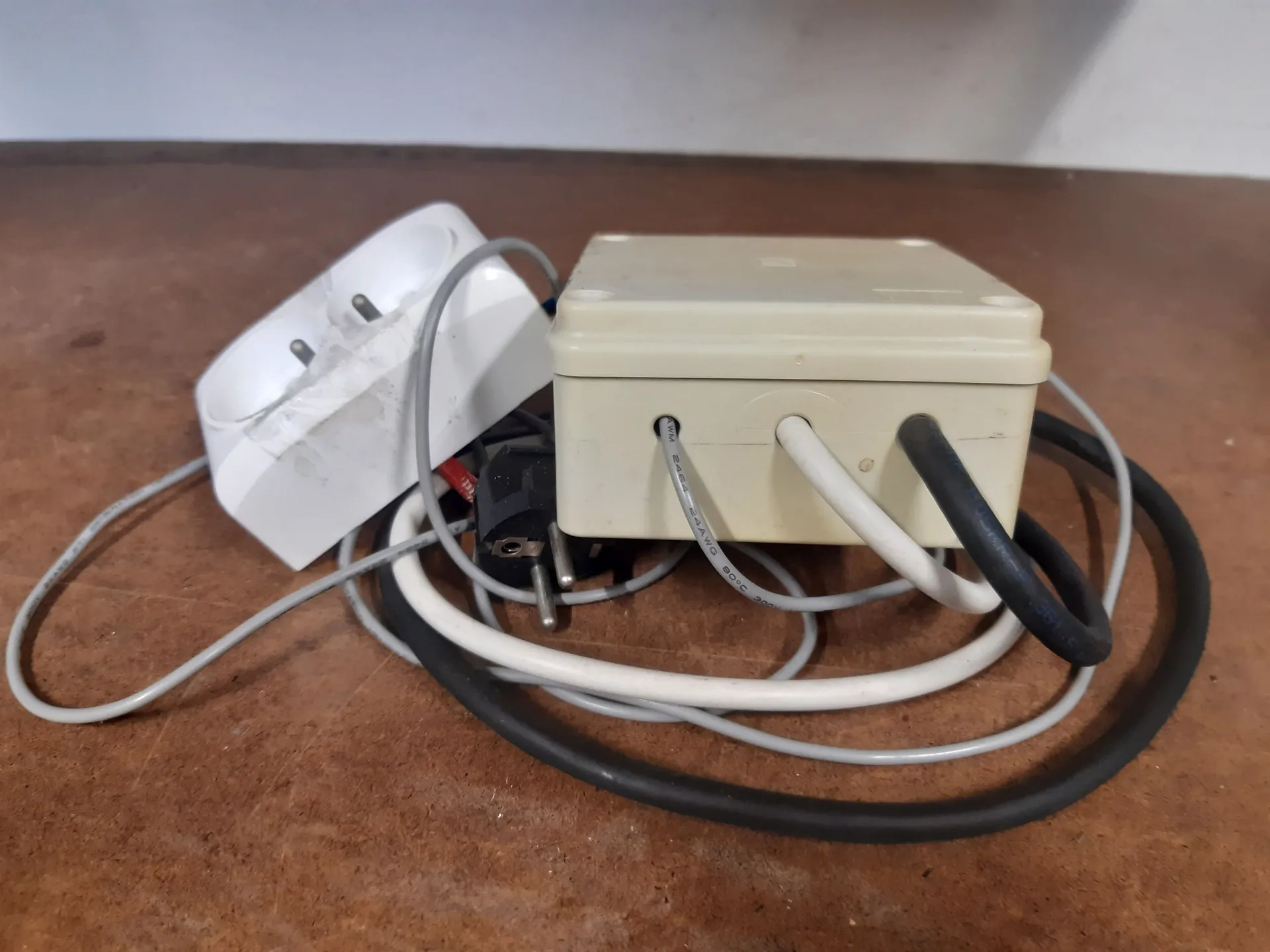 Zestaw elektryczny leżący na drewnianym blacie. W skład zestawu wchodzi biała puszka elektryczna z trzema wyprowadzonymi przewodami (dwoma jasnymi i jednym czarnym), biały przedłużacz z gniazdkiem oraz czarna wtyczka sieciowa. Przewody są splątane wokół puszki.