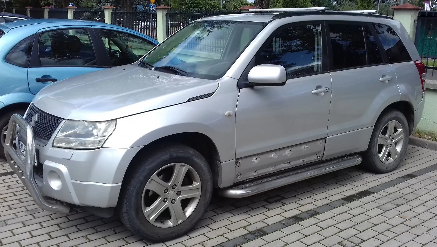 Srebrny samochód typu SUV marki Suzuki zaparkowany na kostce brukowej. Pojazd posiada zamontowany z przodu metalowy zderzak ochronny (tzw. bullbar) oraz stopnie boczne. W tle widoczny jest fragment niebieskiego samochodu oraz ogrodzenie z zielenią.