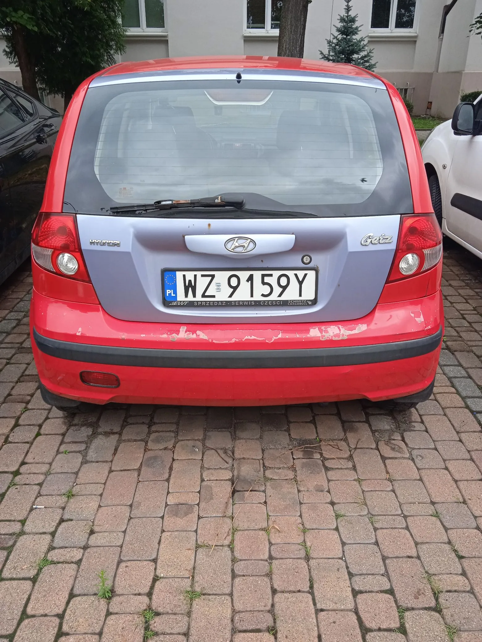 Tylny widok czerwonego samochodu marki Hyundai Getz zaparkowanego na kostce brukowej. Samochód ma srebrną klapę bagażnika z logo Hyundai i napisem 'Getz'. Tablica rejestracyjna to WZ 9159Y. Na tylnym zderzaku widoczne są odpryski lakieru i zarysowania.