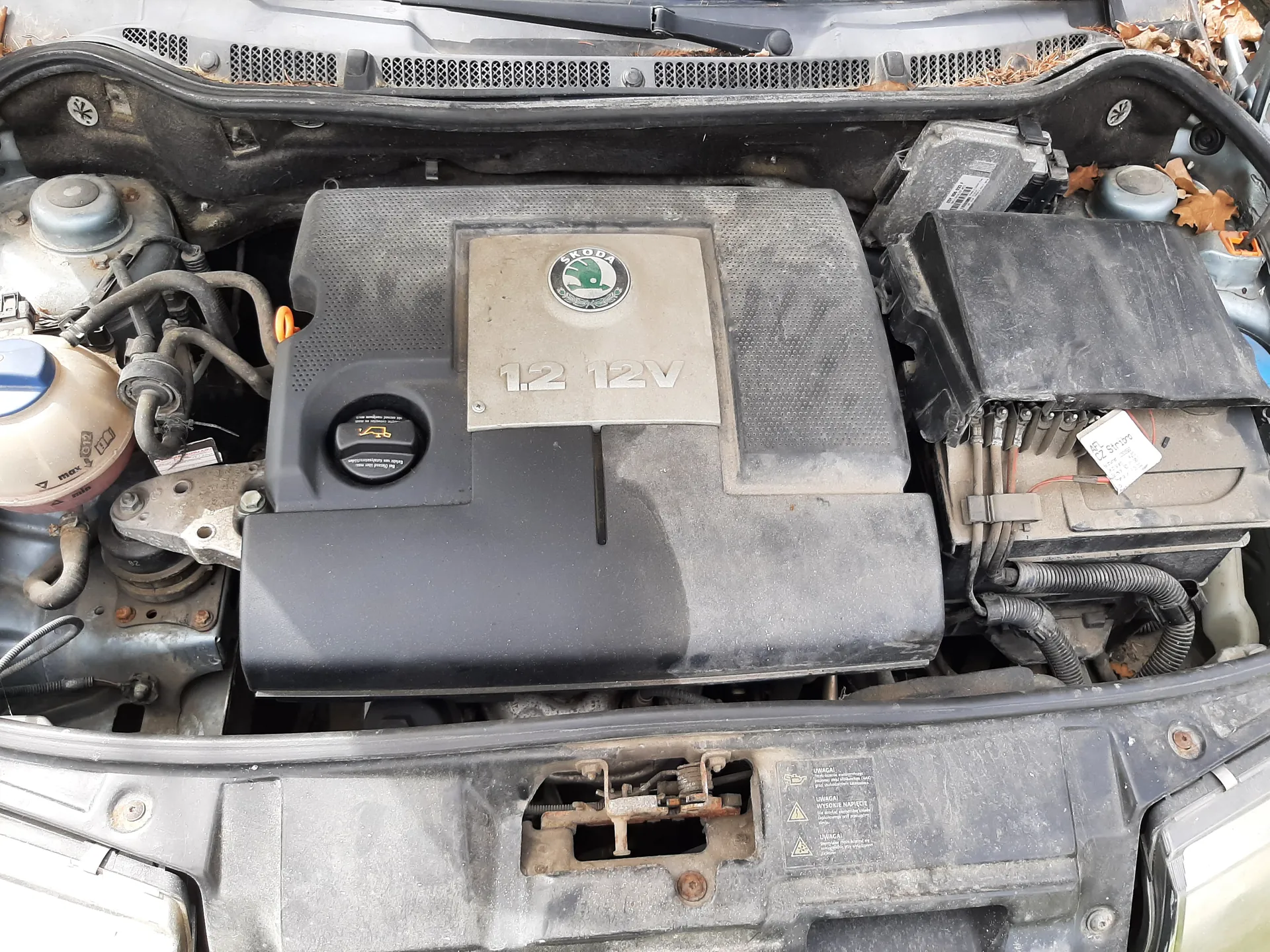 Widok na komorę silnika samochodu marki Škoda. Centralnie umieszczona pokrywa silnika z logo Škoda i napisem „1.2 12V”. Po lewej stronie zbiornik chłodniczy z niebieskim korkiem, przewody i węże. Po prawej stronie czarna skrzynka z białą etykietą oraz suche liście. Komora silnika jest zabrudzona, widoczne elementy mechaniczne i plastikowe osłony.