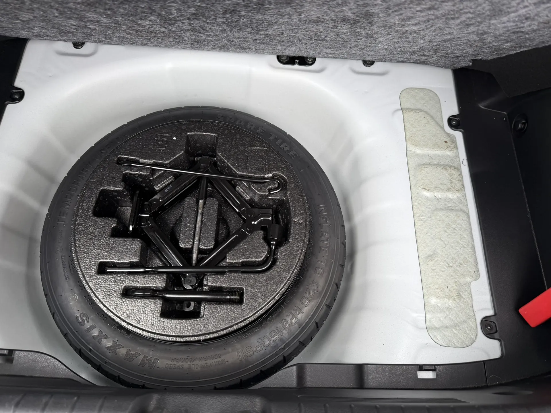 Wnętrze bagażnika samochodu z umieszczonym kołem zapasowym. Czarne koło zapasowe z widocznym napisem 'MAXXIS', 'SPARE TIRE' oraz 'INFLATE TO 420 kPa (60 PSI)'. Na kole leży czarny podnośnik hydrauliczny i narzędzia. Biała obudowa bagażnika z szarą tkaniną na górze oraz czerwonym elementem po prawej stronie.
