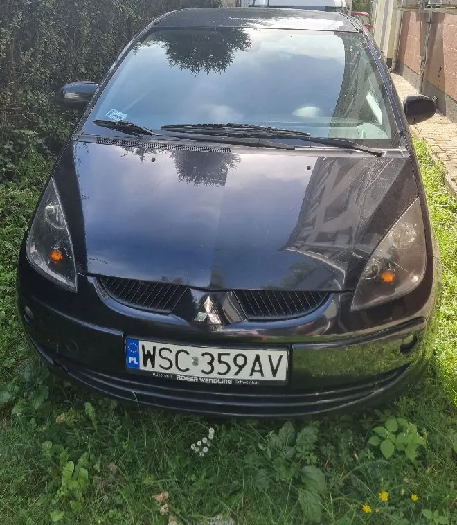 Czarny samochód osobowy marki Mitsubishi widziany od przodu, zaparkowany na trawie. Pojazd posiada polską tablicę rejestracyjną o numerze WSC 359AV. Poniżej tablicy znajduje się napis 'ROGER WENDLING'.