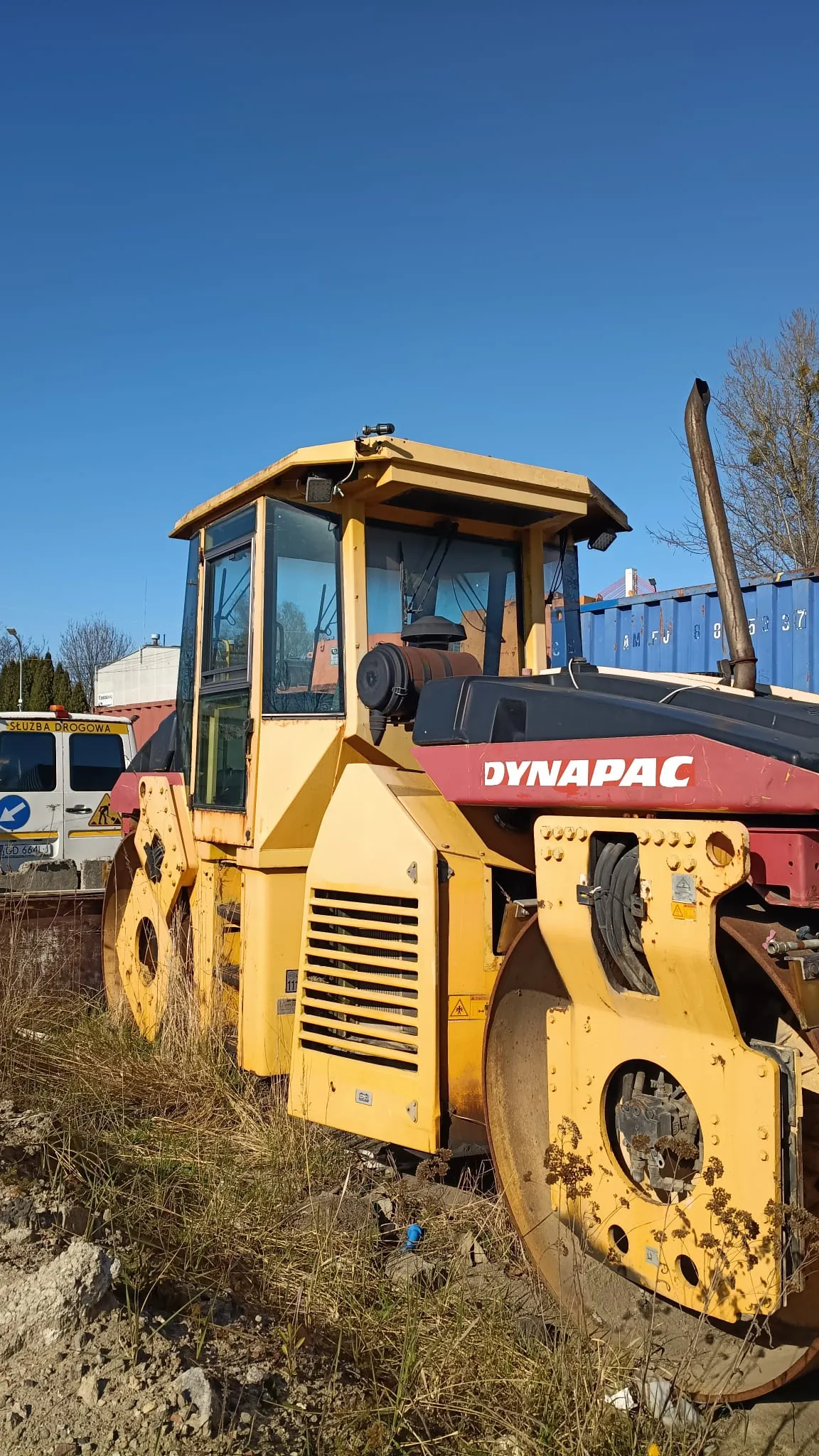 Żółto-czerwony walec drogowy marki Dynapac zaparkowany na nieutwardzonym terenie z trawą. Maszyna posiada żółtą kabinę operatora i czerwony element obudowy z białym napisem „DYNAPAC”. W tle widoczne jest jasnoniebieskie niebo, fragmenty budynków oraz niebieski kontener transportowy.