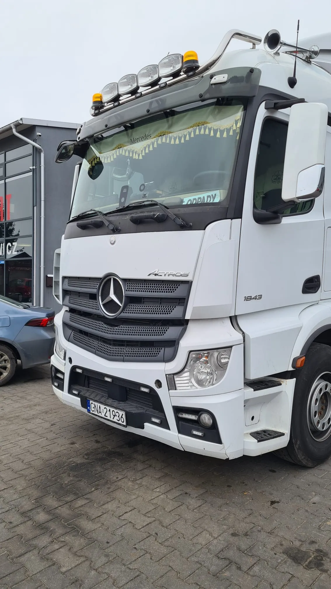 Biały ciągnik siodłowy marki Mercedes-Benz Actros 1843. Pojazd posiada dodatkowe oświetlenie na dachu, tabliczkę z napisem 'ODPADY' za przednią szybą oraz polską tablicę rejestracyjną GNA 21936. Ciągnik stoi na brukowanym placu obok szarego budynku i fragmentu niebieskiego samochodu osobowego.