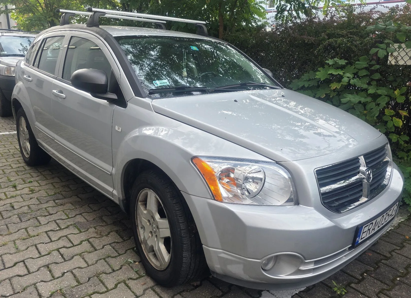 Srebrny samochód marki Dodge Caliber zaparkowany na kostce brukowej. Pojazd posiada czarne lusterka boczne oraz srebrny bagażnik dachowy. W tle widoczne są zielone krzewy i drzewa.
