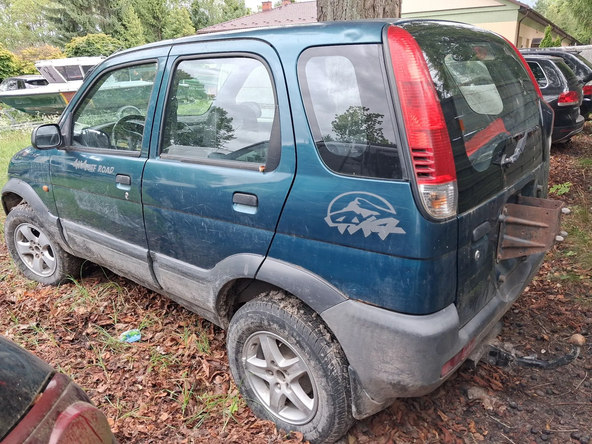 Niebieski samochód typu hatchback zaparkowany na ziemi pokrytej suchymi liśćmi i trawą. Pojazd jest zabrudzony, posiada szare plastikowe osłony nadkoli i progi. Na tylnym błotniku znajduje się szara naklejka z motywem gór i napisem 4x4, a na przednich drzwiach napis OFF ROAD. Samochód jest widoczny z profilu od strony pasażera, z tyłu brakuje klapy bagażnika. W tle widoczne są inne samochody, łódź oraz fragmenty budynków i drzew.