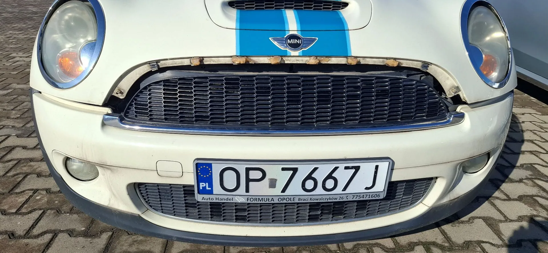 Zbliżenie na przód białego samochodu marki Mini. Na masce widoczne są dwa niebieskie pasy oraz logo Mini. Samochód posiada czarny grill z chromowaną obramowaniem, nad którym widoczne są ślady korozji. Na zderzaku zamontowana jest polska tablica rejestracyjna o numerze OP 7667J. Poniżej numeru rejestracyjnego znajduje się napis: 'Auto Handel FORMUŁA OPOLE Braci Kowalczyków 26 775471606'.