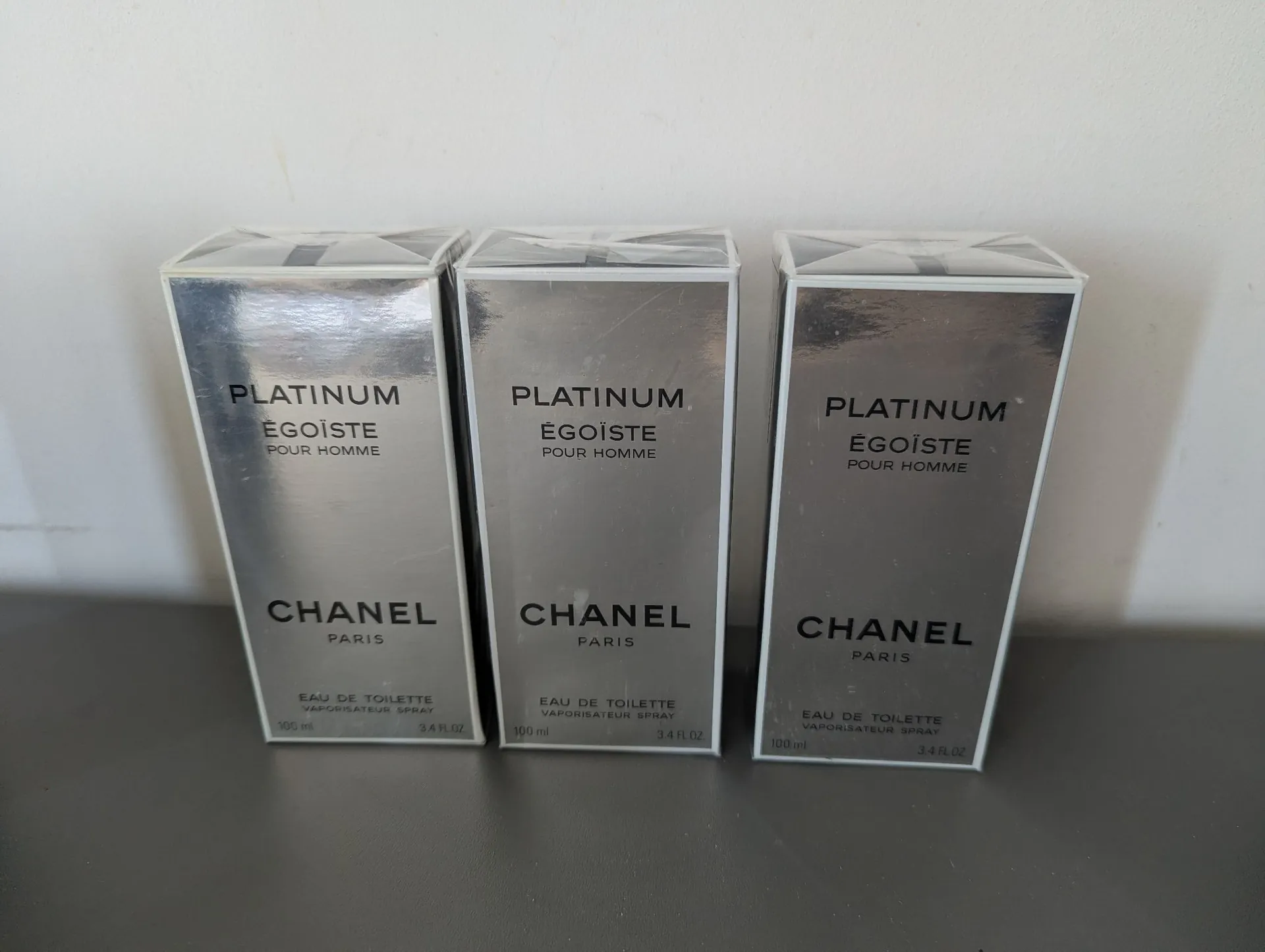 Trzy identyczne, srebrne pudełka perfum Chanel Platinum Égoïste Pour Homme ustawione obok siebie na ciemnej powierzchni na tle białej ściany. Na każdym pudełku widnieją napisy: 'PLATINUM ÉGOÏSTE POUR HOMME', 'CHANEL PARIS' oraz 'EAU DE TOILETTE VAPORISATEUR SPRAY 100 ml 3.4 FL.OZ'.
