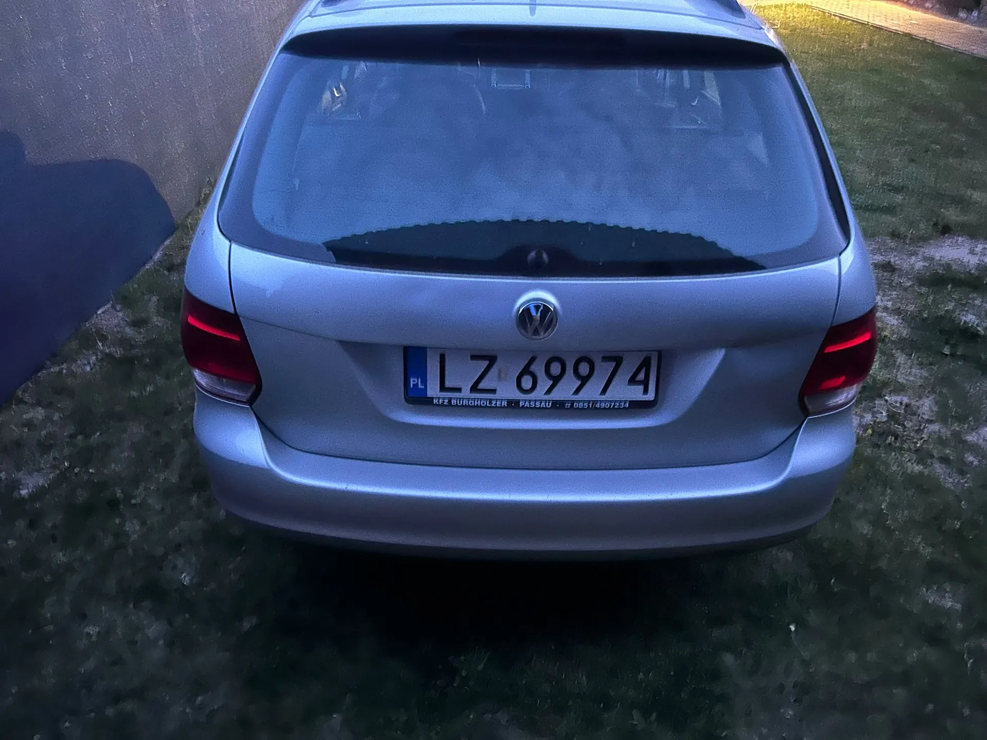 Tylny widok srebrnego samochodu marki Volkswagen. Na klapie bagażnika znajduje się logo producenta oraz polska tablica rejestracyjna o numerze LZ 69974. Poniżej tablicy widnieje napis: KFZ BURGHOLZER - PASSAU - 0851/4901234. Samochód stoi na trawie obok betonowej ściany.