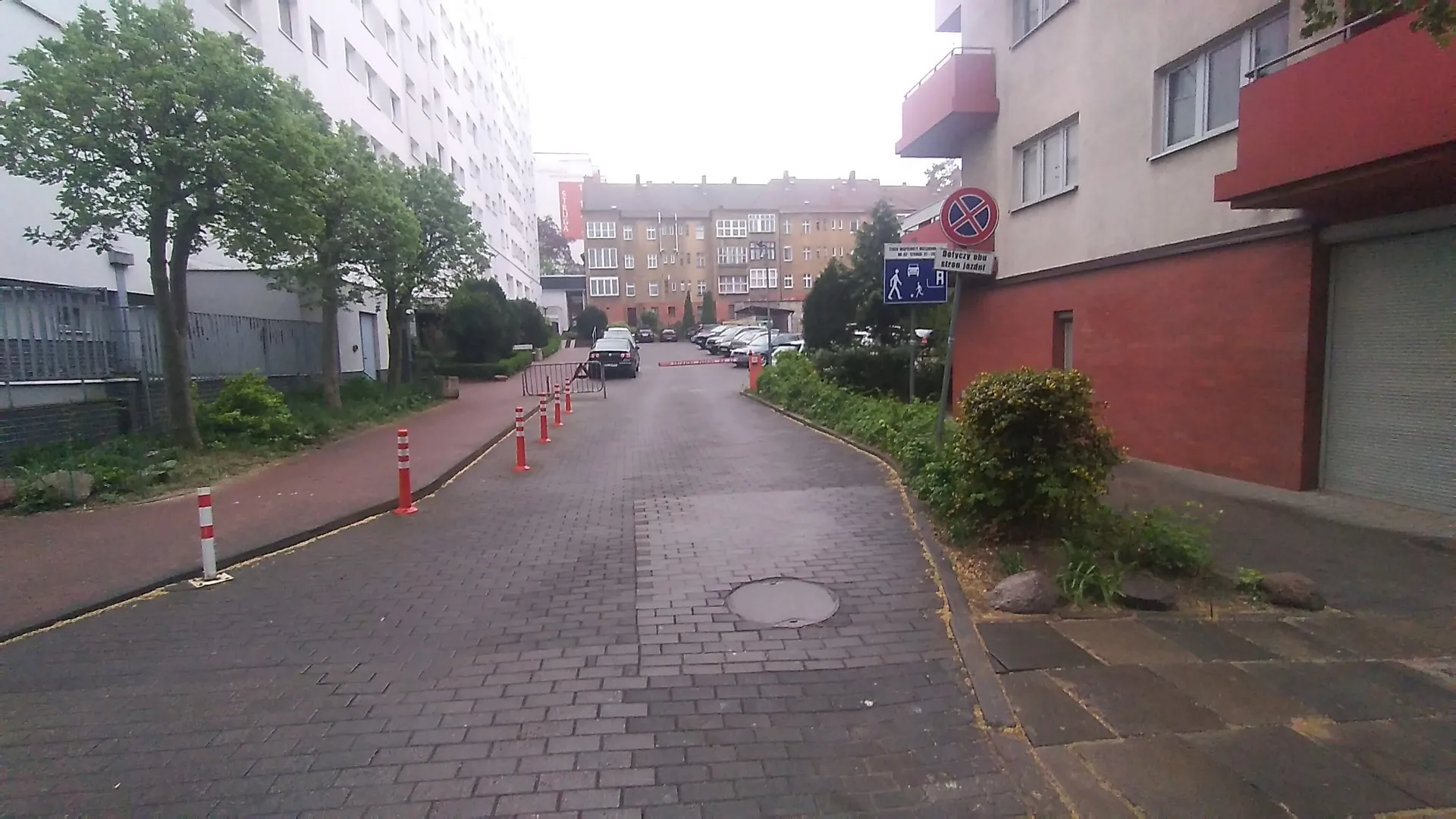 Widok na osiedlową drogę wyłożoną szarą kostką brukową, prowadzącą między blokami mieszkalnymi. Po lewej stronie znajduje się biały budynek z rzędem drzew i niskim ogrodzeniem, a po prawej czerwony budynek z garażem. Wzdłuż drogi ustawione są biało-czerwone pachołki drogowe. W oddali widoczne są zaparkowane samochody oraz blok mieszkalny. Po prawej stronie znajduje się znak zakazu zatrzymywania się oraz niebieska tabliczka informacyjna z piktogramem pieszego i tekstem.
