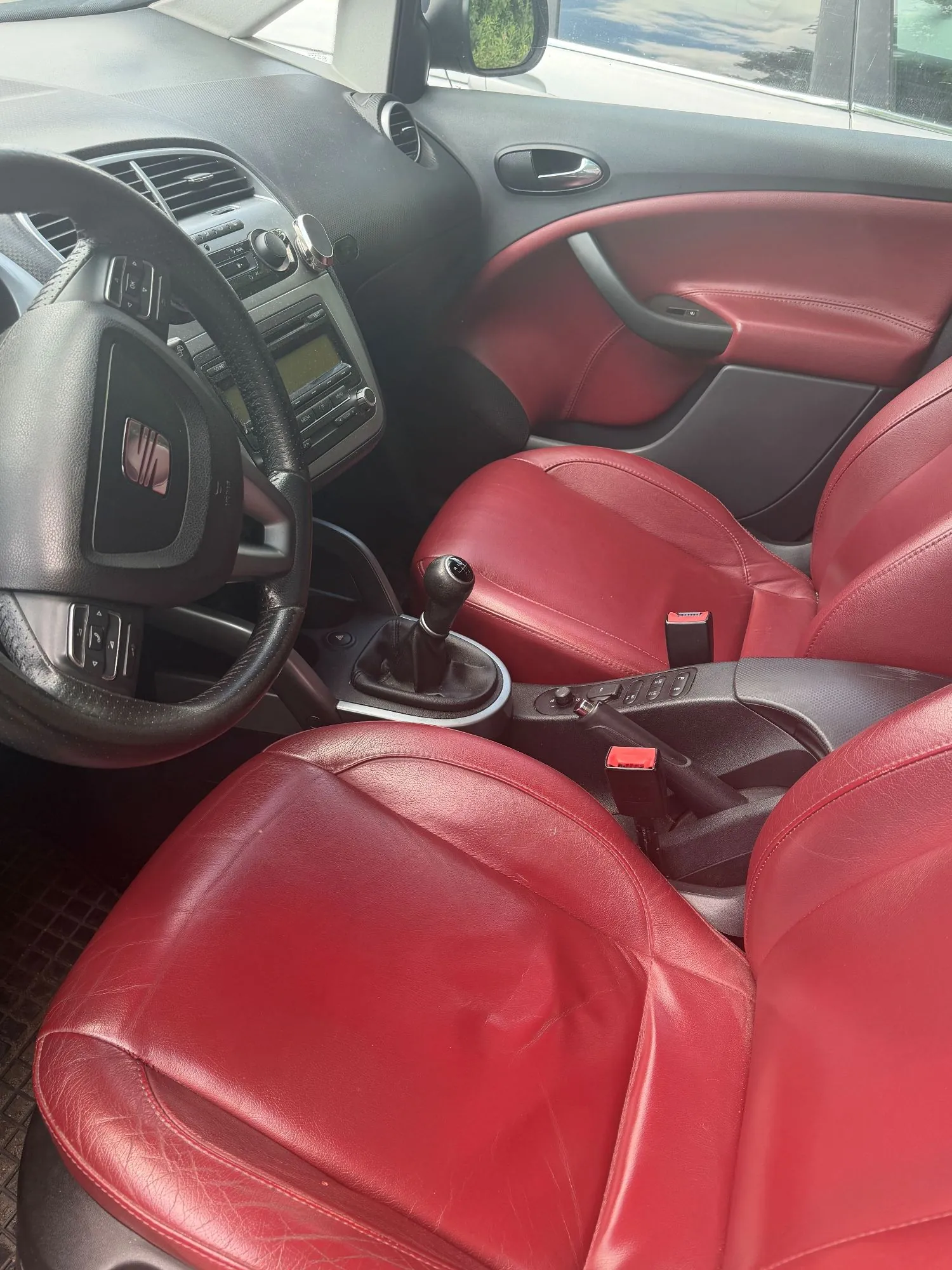 Wnętrze samochodu z czerwonymi, skórzanymi fotelami. Widoczna jest kierownica z logo marki SEAT, deska rozdzielcza z radiem, lewarek zmiany biegów oraz czerwona tapicerka drzwi kierowcy.