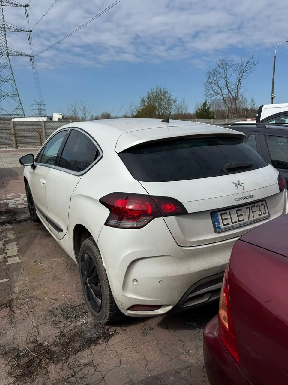 Biały samochód Citroën z tyłu, z widoczną tablicą rejestracyjną PL ELE 7F33. Pojazd zaparkowany na brukowanej powierzchni, częściowo widoczne są inne samochody (czerwony i czarny). W tle betonowy płot, wysokie drzewa bez liści, linie energetyczne z masztami oraz niebo z rozproszonymi chmurami.