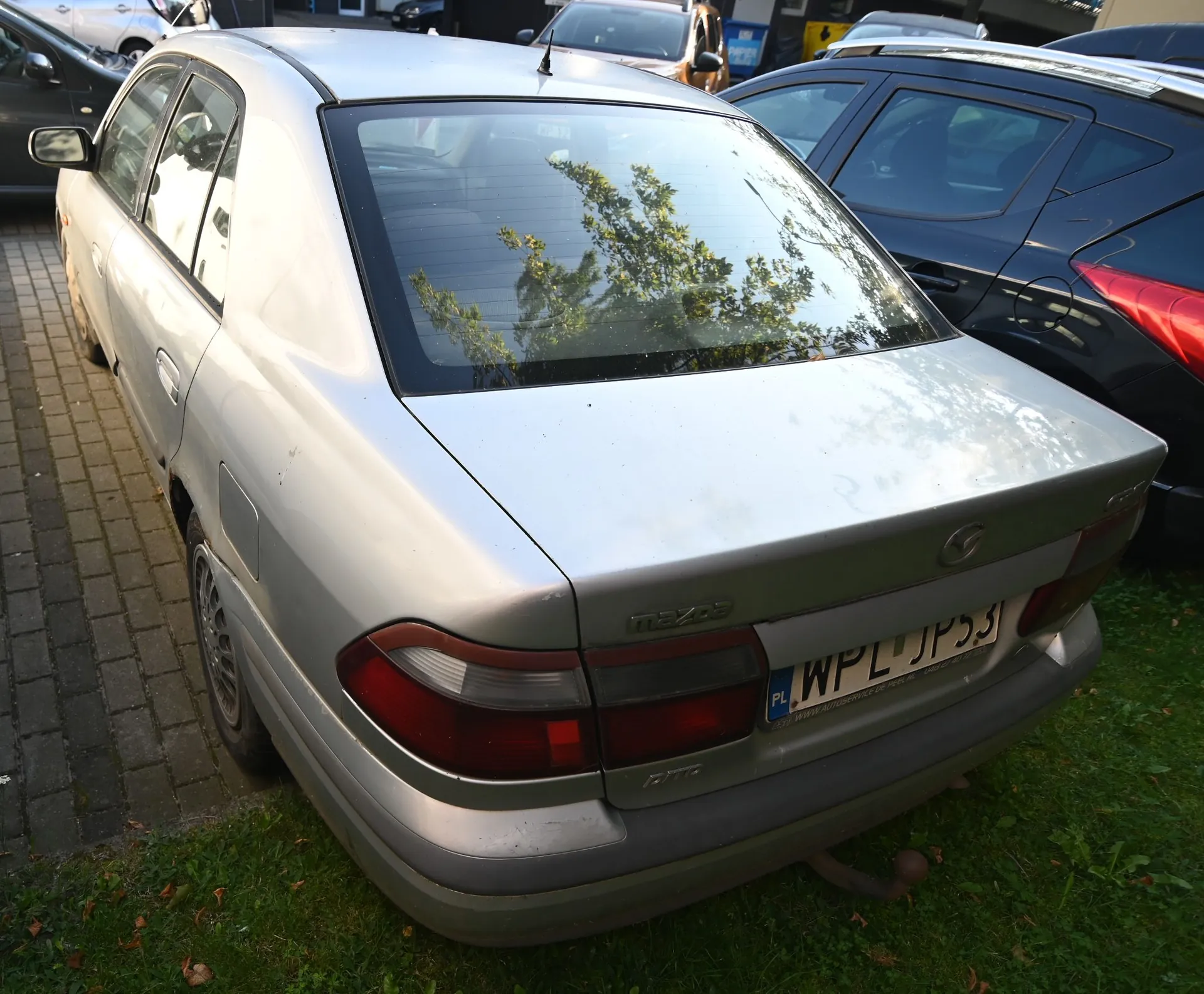 Srebrny samochód marki Mazda widziany od tyłu i z boku, zaparkowany na kostce brukowej obok trawnika. Na klapie bagażnika widnieje napis 'mazda' oraz tablica rejestracyjna o numerze 'WPL JP53'. W tle widoczne są inne zaparkowane samochody, w tym czarny pojazd po prawej stronie.