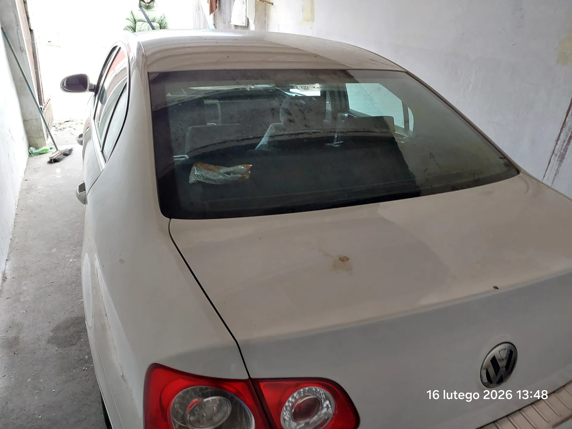 Biały samochód marki Volkswagen, widok od tyłu, zaparkowany w wąskim korytarzu z białymi ścianami. Widoczne czerwone światła tylnego, logo VW na bagażniku oraz przedmioty wewnątrz przez tylne szyby. W tle osoba w zielonej kurtce, a na podłodze po lewej stronie narzędzia i zielona butelka. W prawym dolnym rogu obrazu widoczny tekst: 16 lutego 2026 13:48.