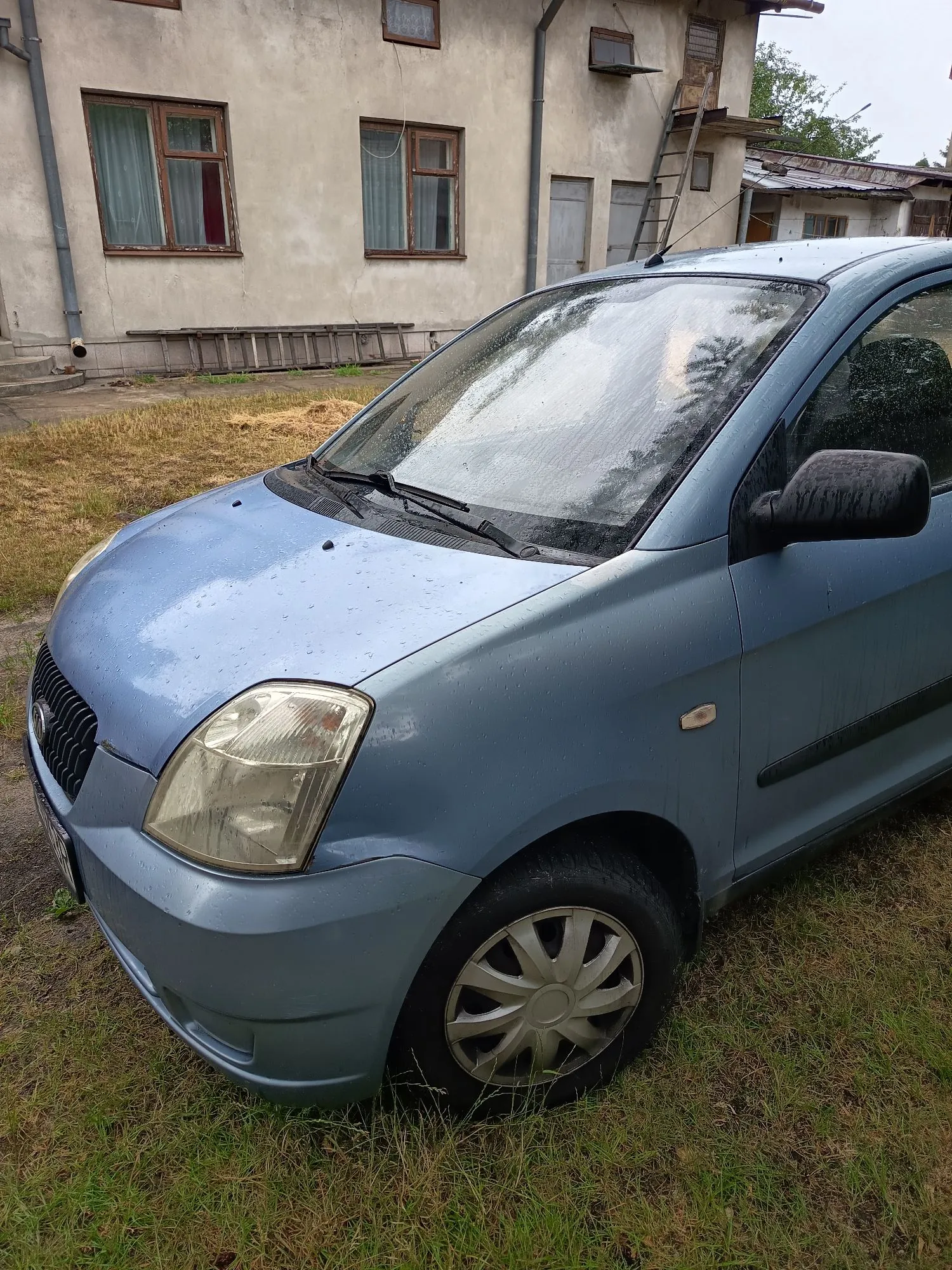 Jasnoniebieski samochód osobowy zaparkowany na trawiastym podłożu przed starym, jasnym budynkiem z widocznymi oknami i drabiną opartą o ścianę. Auto jest mokre od deszczu, widoczne są krople wody na masce i przedniej szybie.