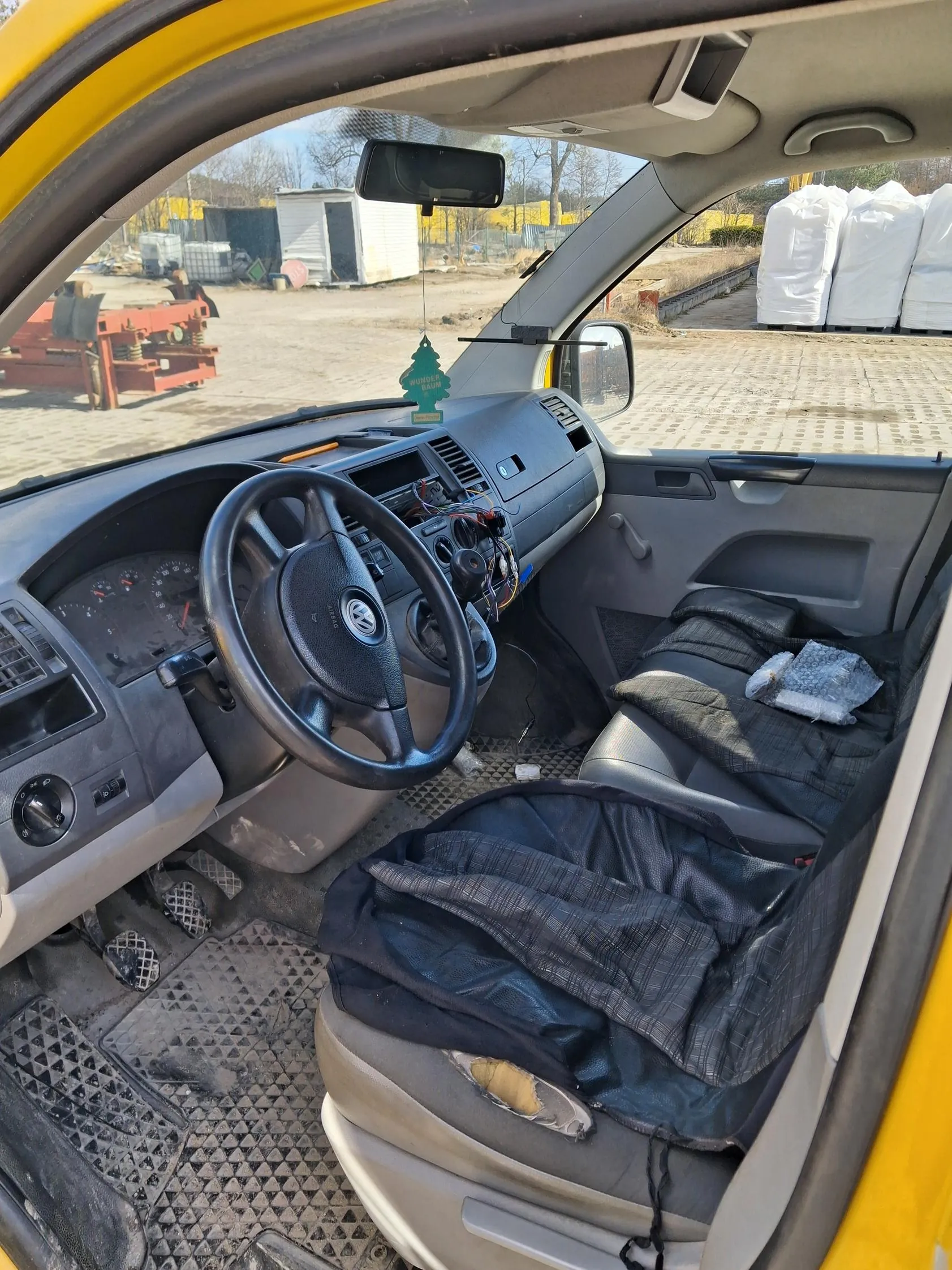 Wnętrze żółtego pojazdu z widoczną kierownicą marki Volkswagen. Deska rozdzielcza z odsłoniętymi przewodami, zielony odświeżacz powietrza z napisem "WUNDER-BAUM" wiszący na lusterku wstecznym. Siedzenia pokryte ciemnym materiałem, brudne dywaniki podłogowe. Poza pojazdem przemysłowy teren z czerwonymi urządzeniami, białymi budynkami i białymi owiniętymi przedmiotami.