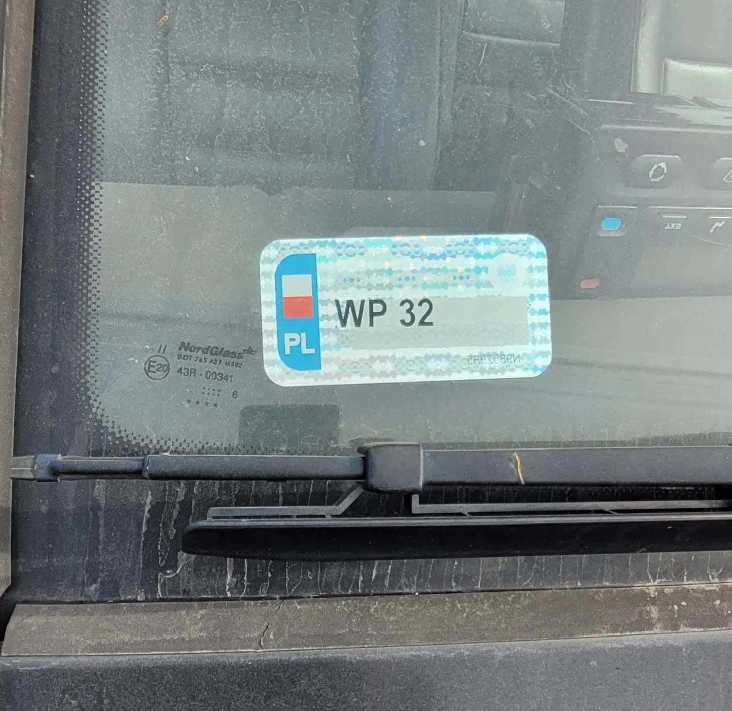 Zbliżenie na tylną szybę samochodu, na której naklejona jest niebiesko-biała naklejka z napisem 'PL' oraz kodem 'WP 32'. Poniżej naklejki widoczna jest czarna wycieraczka szyby. Po lewej stronie znajduje się oznaczenie producenta szkła 'NordGlass' wraz z numerami seryjnymi i certyfikatem E20.