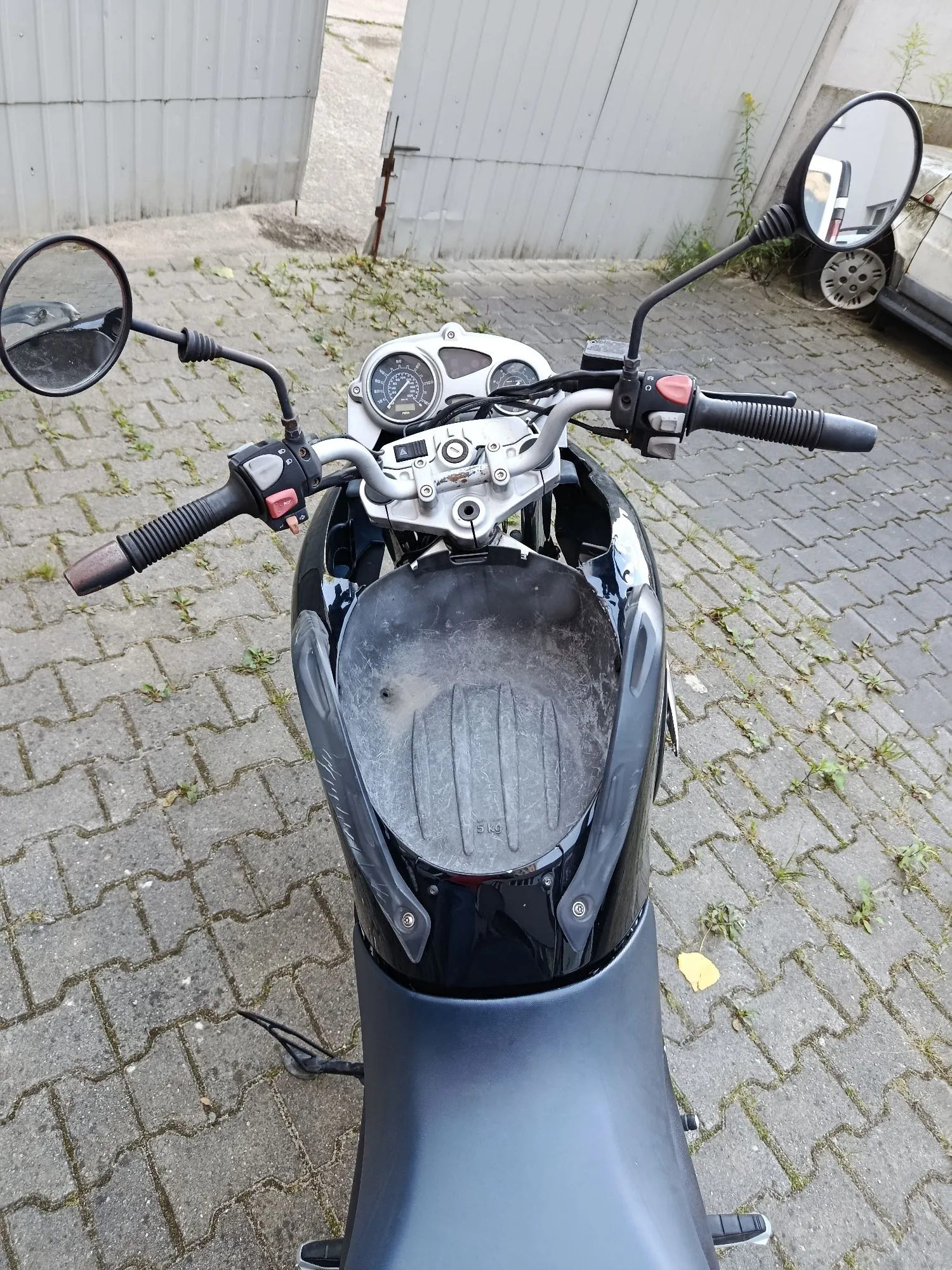 Widok z perspektywy kierowcy na czarny skuter zaparkowany na kostce brukowej. Widoczny jest kierownica z manetkami, lusterka boczne, zestaw wskaźników z prędkościomierzem oraz schowek przed kolanami. W tle znajduje się jasny płot i fragment srebrnego samochodu.