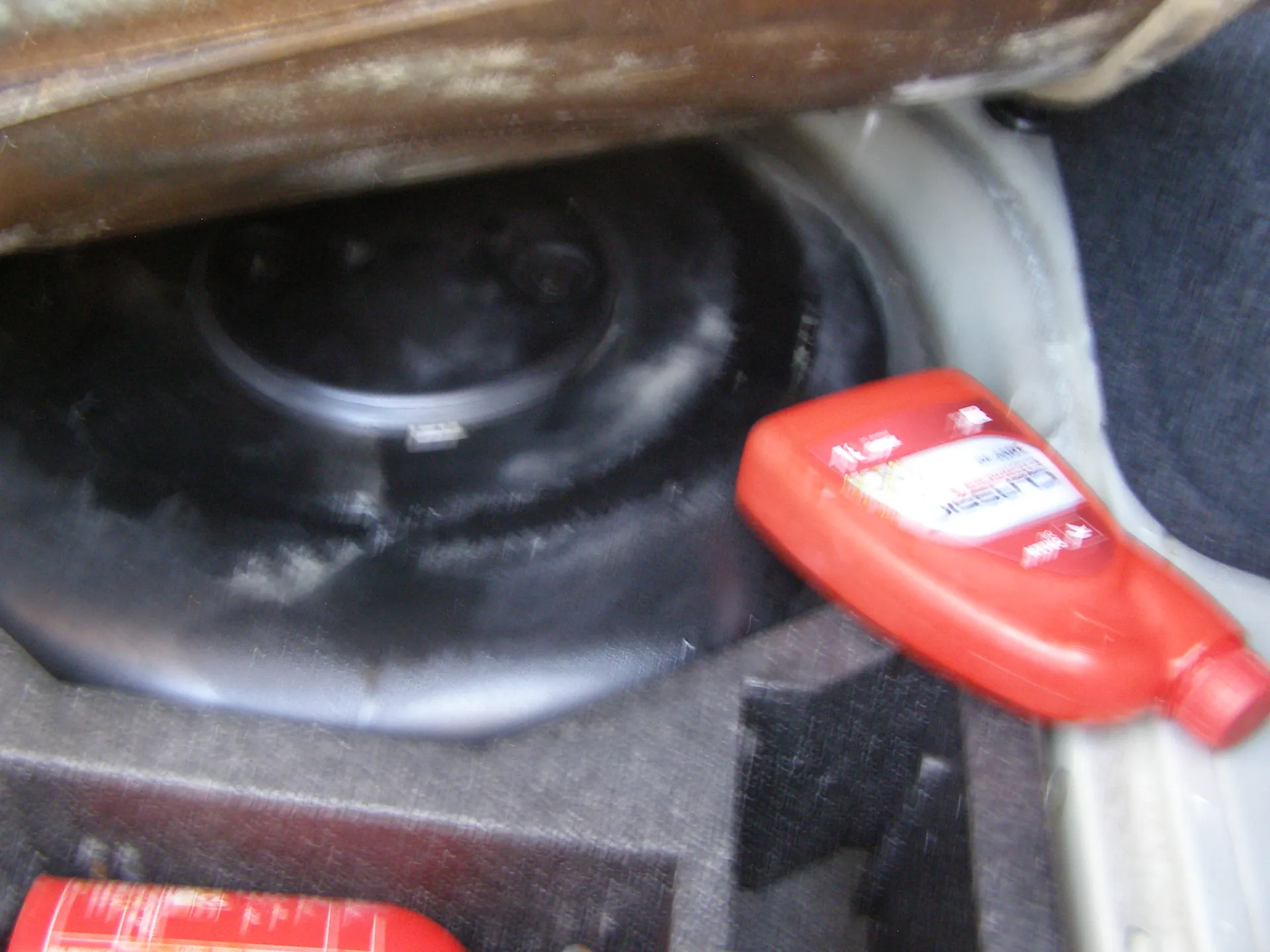 Wnętrze bagażnika samochodu z czarnym kołem zapasowym w osłonie. Obok koła znajduje się czerwona butelka z etykietą zawierającą czytelny napis 'LIQUID' oraz częściowo widoczny czerwony pojemnik. Widoczne są również elementy wnętrza samochodu, takie jak tkanina i metalowe części.