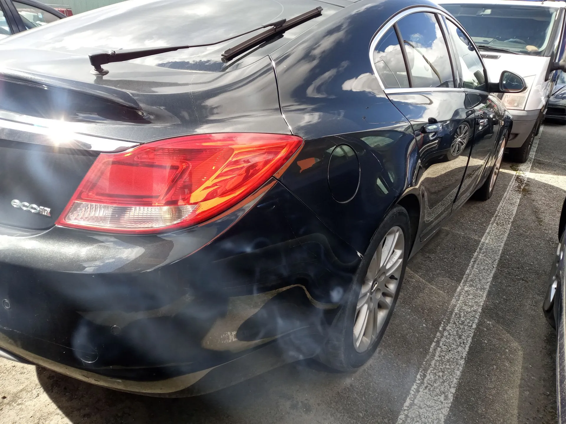 Tylna prawa część ciemnogranatowego samochodu osobowego zaparkowanego na asfaltowym parkingu. Widoczny jest tylny reflektor, klapka wlewu paliwa oraz tylne koło z felgą aluminiową. W tle widoczny jest fragment innego, białego samochodu.
