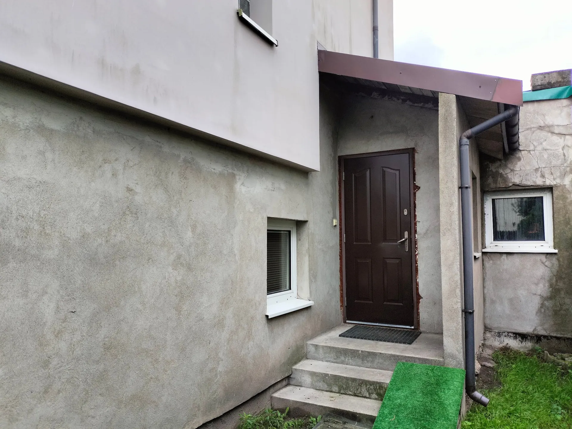 Zdjęcie fragmentu budynku z szarymi, tynkowanymi ścianami. Widoczne są brązowe drzwi wejściowe z czterema panelami, do których prowadzą trzy betonowe stopnie. Obok drzwi znajduje się małe, kwadratowe okno z białą ramą. Po prawej stronie widoczna jest szara rura spustowa oraz kolejne małe okno w ścianie. Przed schodami leży zielona wycieraczka z sztucznej trawy.