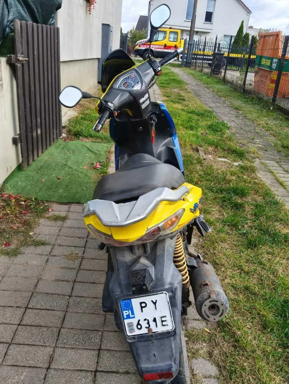 Widok od tyłu na niebiesko-żółty skuter zaparkowany na kostce brukowej obok trawnika. Skuter posiada tablicę rejestracyjną o numerze PY 631E z oznaczeniem PL. W tle widoczny jest żółty samochód dostawczy z napisem POMOC oraz fragmenty zabudowań i ogrodzenia.