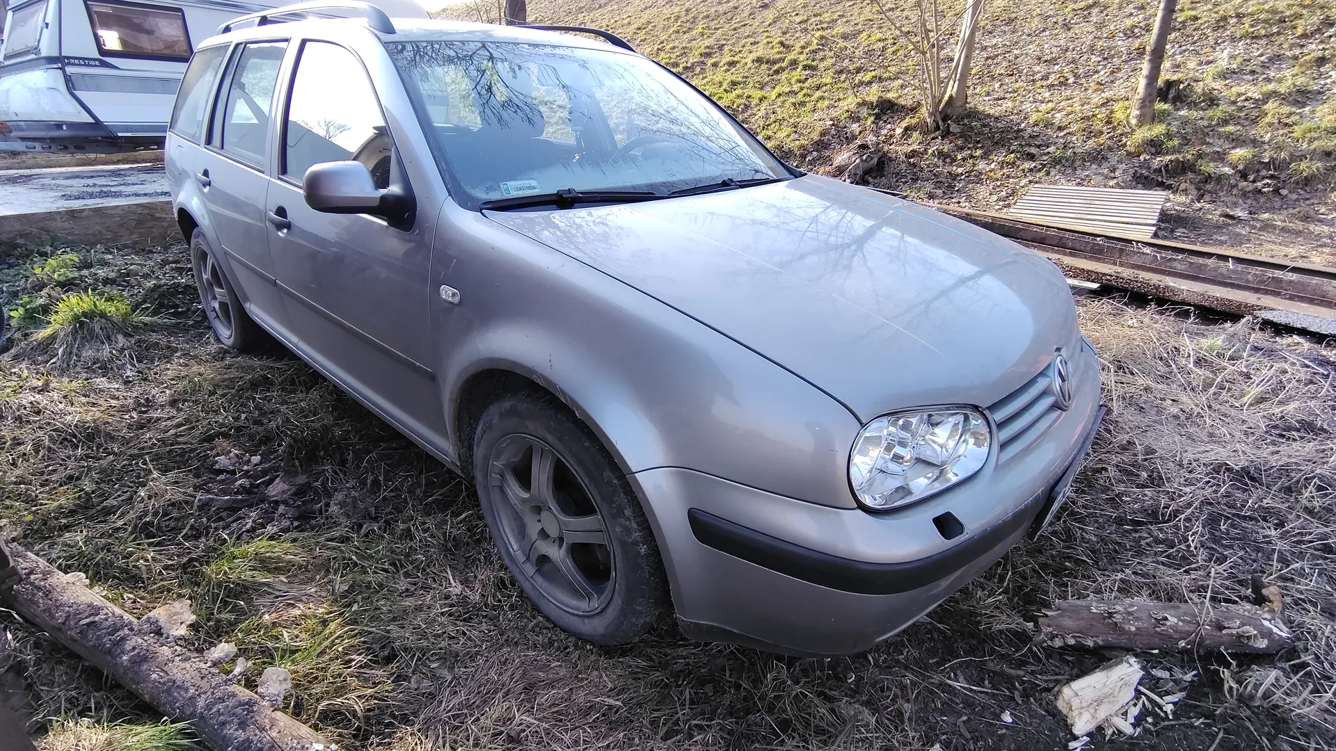 Srebrny samochód typu kombi marki Volkswagen zaparkowany na trawiastym i błotnistym terenie. W tle widoczna przyczepa z napisem 'Prestige', tor kolejowy oraz pnie drzew. Na ziemi leżą kawałki drewna i sucha trawa.