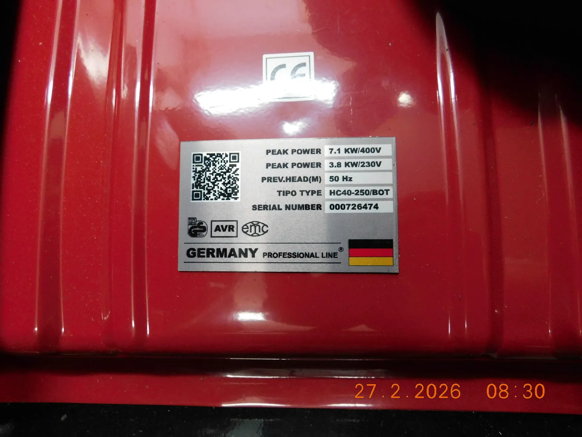 Zbliżenie na czerwoną, metalową obudowę urządzenia z przytwierdzoną srebrną tabliczką znamionową. Na tabliczce znajduje się kod QR oraz następujące dane techniczne: PEAK POWER 7.1 KW/400V, PEAK POWER 3.8 KW/230V, PREV.HEAD(M) 50 Hz, TIPO TYPE HC40-250/BOT, SERIAL NUMBER 000726474. Poniżej widnieją oznaczenia AVR, emc oraz napis GERMANY PROFESSIONAL LINE obok flagi Niemiec. W prawym dolnym rogu zdjęcia widoczna jest data i godzina: 27.2.2026 08:30.