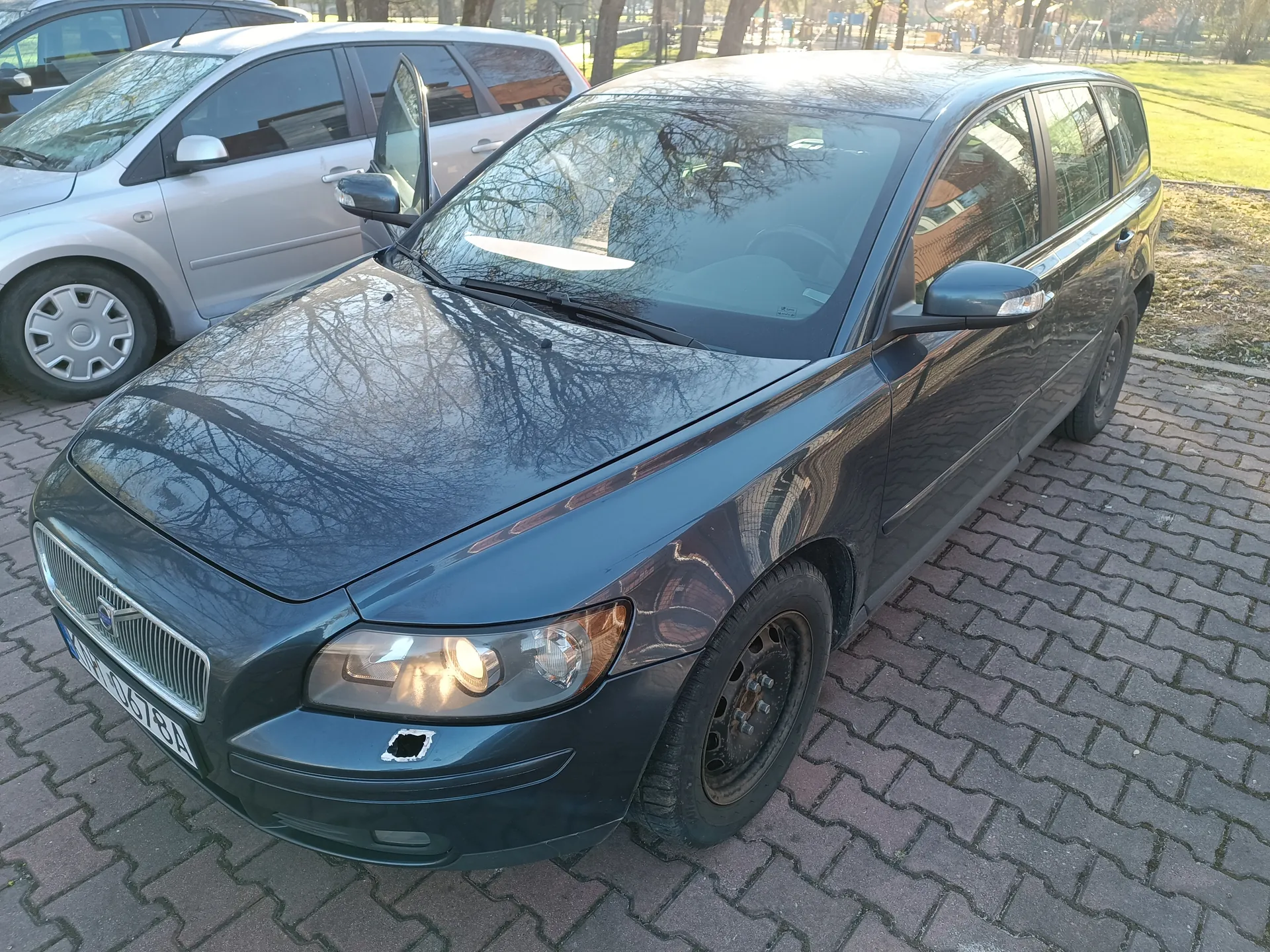 Ciemnoszary samochód osobowy typu kombi marki Volvo zaparkowany na brukowanej powierzchni. Widoczne są włączony lewy reflektor, uszkodzenie na przednim zderzaku oraz tablica rejestracyjna z białym tłem i czarnym napisem AP 0678A. Za nim stoi srebrny samochód z otwartą drzwiami kierowcy. Tło stanowią drzewa, trawnik i budynki w oddali. Słoneczne oświetlenie rzuca cienie na powierzchnię.