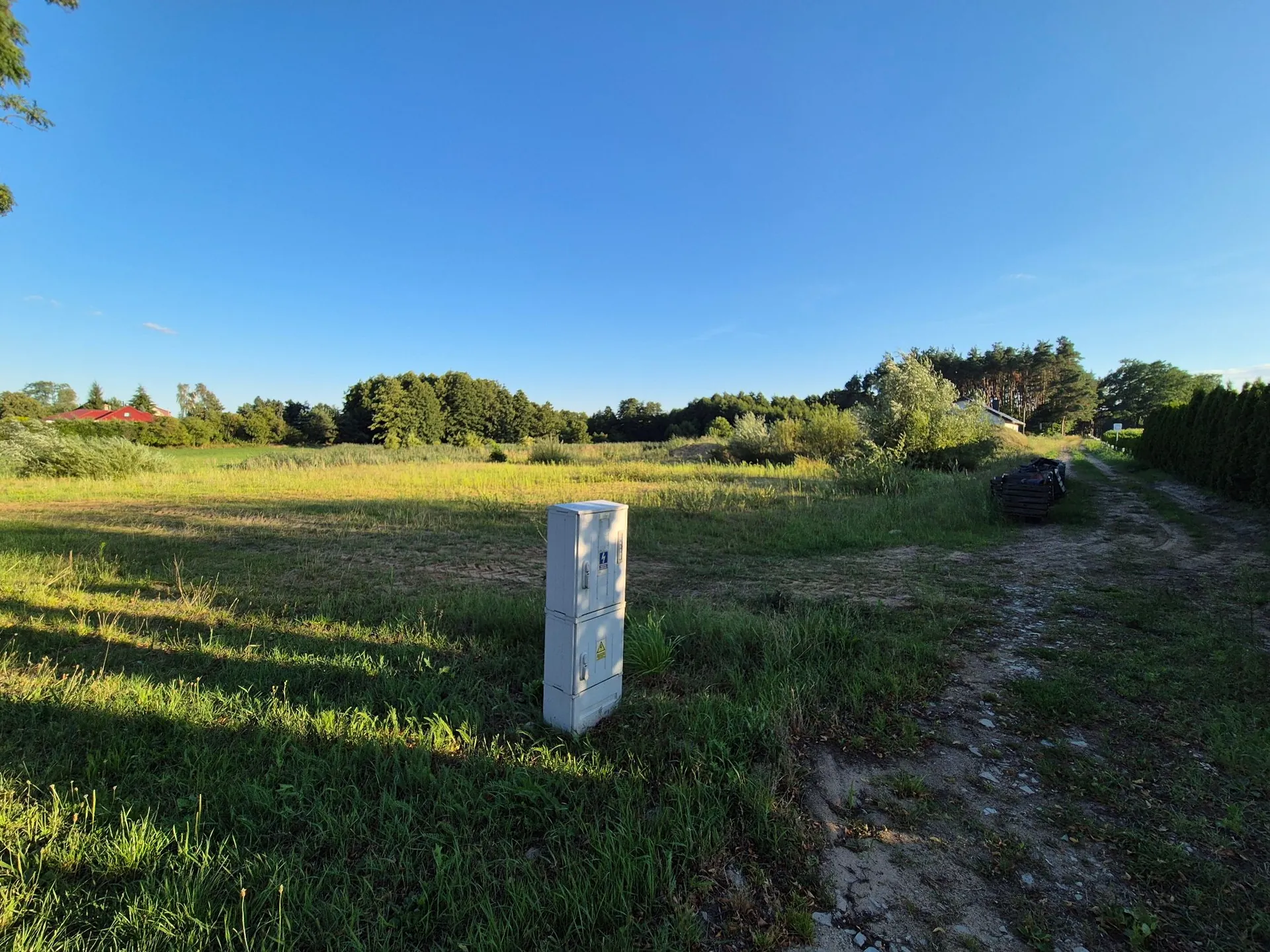 Szeroki kadr przedstawiający otwartą, trawiaste pole pod bezchmurnym, niebieskim niebem. Na pierwszym planie, nieco po lewej stronie, stoi biała, metalowa szafka elektryczna z widocznym żółtym trójkątnym znakiem ostrzegawczym. Po prawej stronie znajduje się nieutwardzona, piaszczysta droga prowadząca w głąb obrazu, wzdłuż której widać ciemne materiały budowlane lub rury. W tle rozciąga się linia zielonych drzew i krzewów, a po lewej stronie widoczny jest fragment czerwonego dachu budynku.