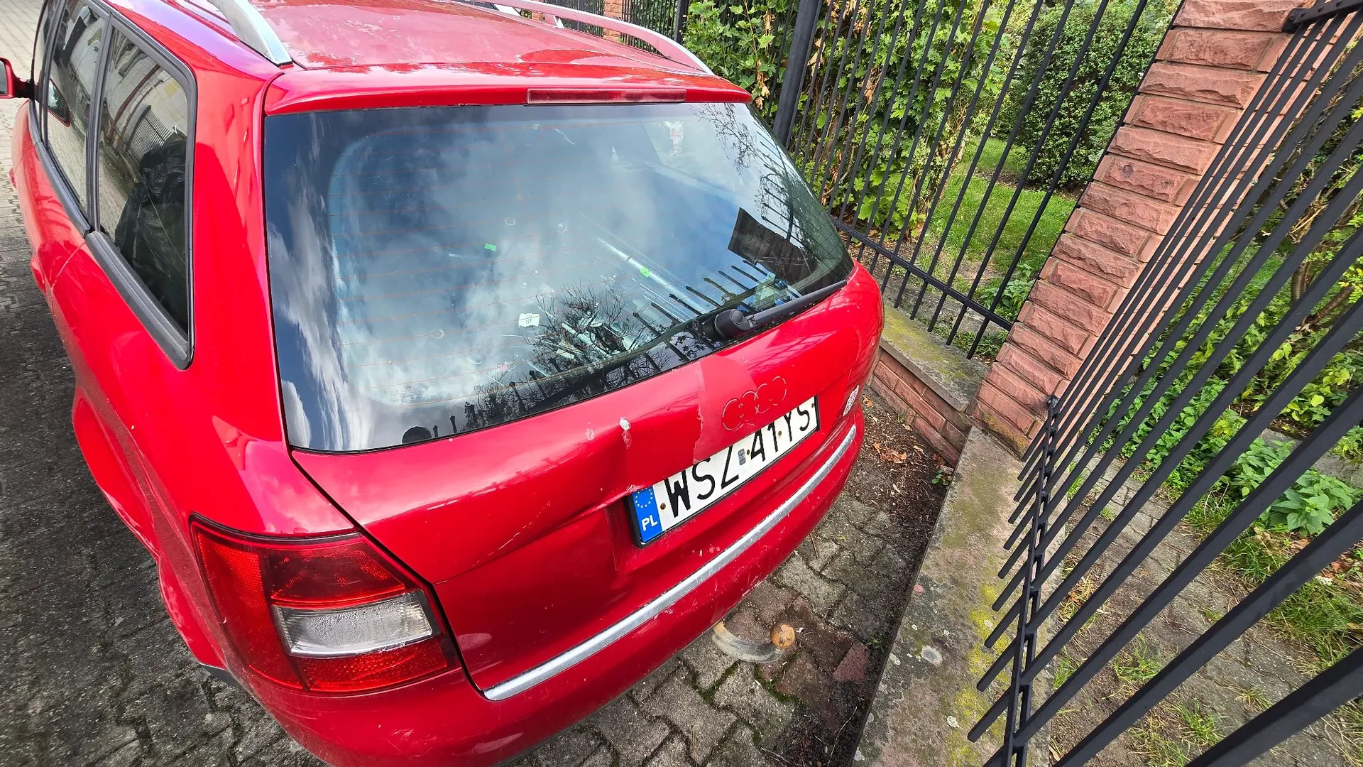 Tylna część czerwonego samochodu typu kombi zaparkowanego na kostce brukowej obok czarnego metalowego ogrodzenia i ceglanego słupka. Na klapie bagażnika widoczna jest tablica rejestracyjna o numerze WSZ 411TS.