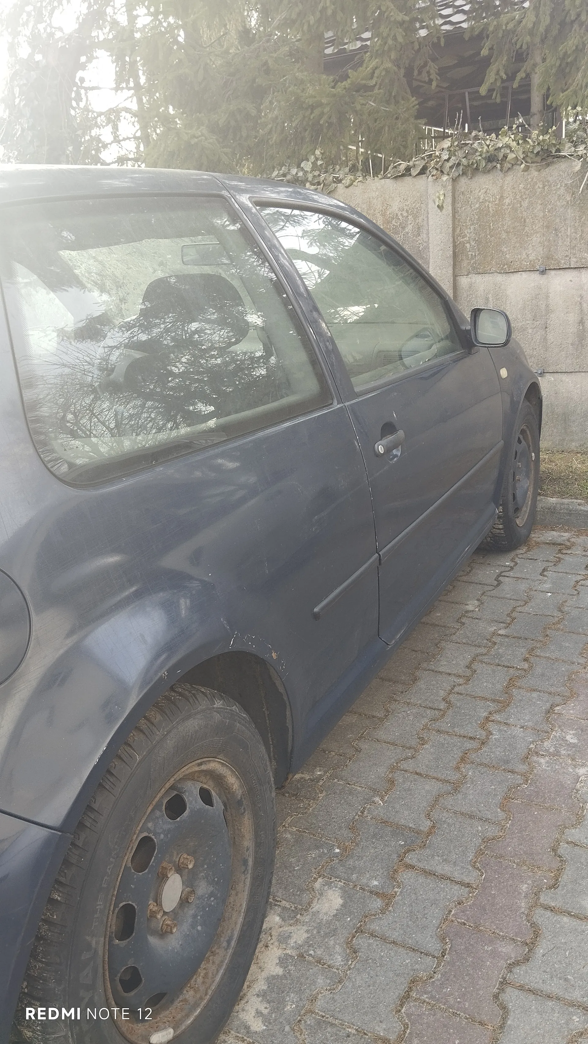 Niebieski samochód osobowy zaparkowany na brukowanej powierzchni z kamiennymi płytami. Samochód ma widoczne ślady zużycia, w tym rdzę na felgach, szczególnie na przednim kole. Po prawej stronie betonowa ściana z roślinnością na szczycie. W tle drzewa i fragment budynku z dachem. Na dole zdjęcia widoczny napis 'REDMI NOTE 12'.
