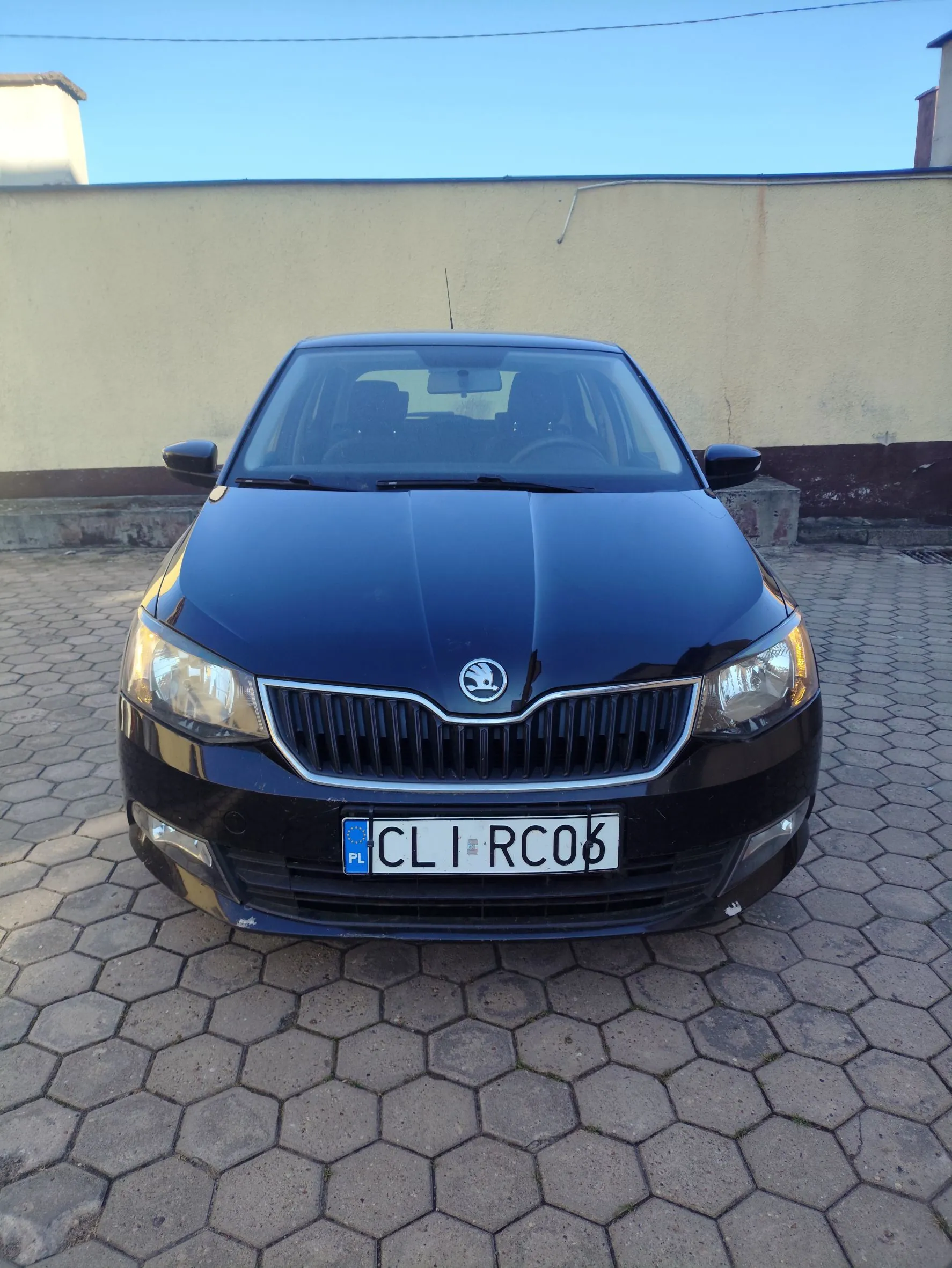 Czarny samochód marki Skoda zaparkowany przodem do widza na kostce brukowej o sześciokątnym kształcie. Pojazd posiada polską tablicę rejestracyjną o numerze CLI RC06. W tle widoczna jest jasna ściana budynku pod błękitnym niebem.