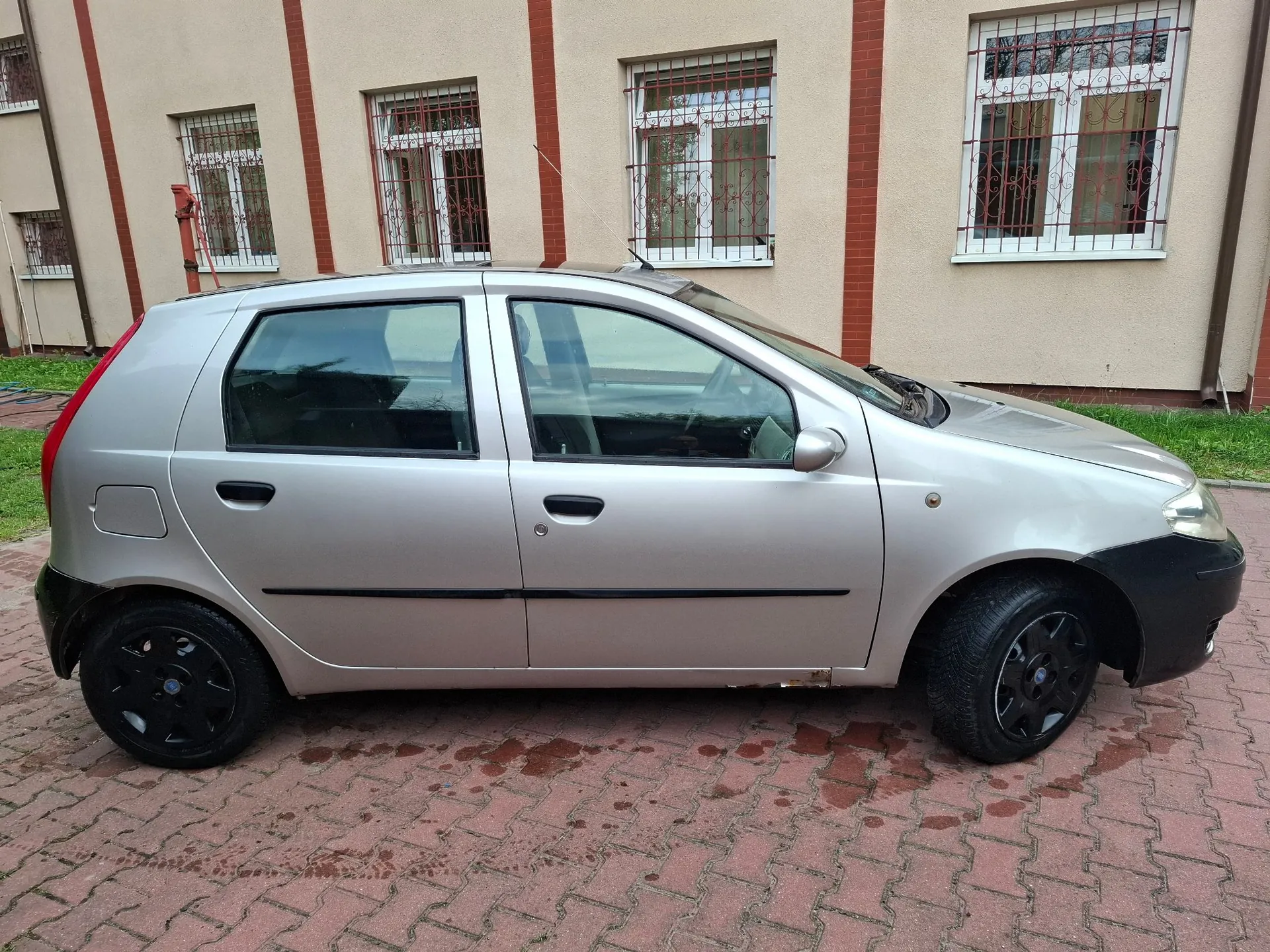 Srebrny samochód osobowy w nadwoziu hatchback zaparkowany bokiem na kostce brukowej przed jasnym budynkiem z oknami zabezpieczonymi kratami.
