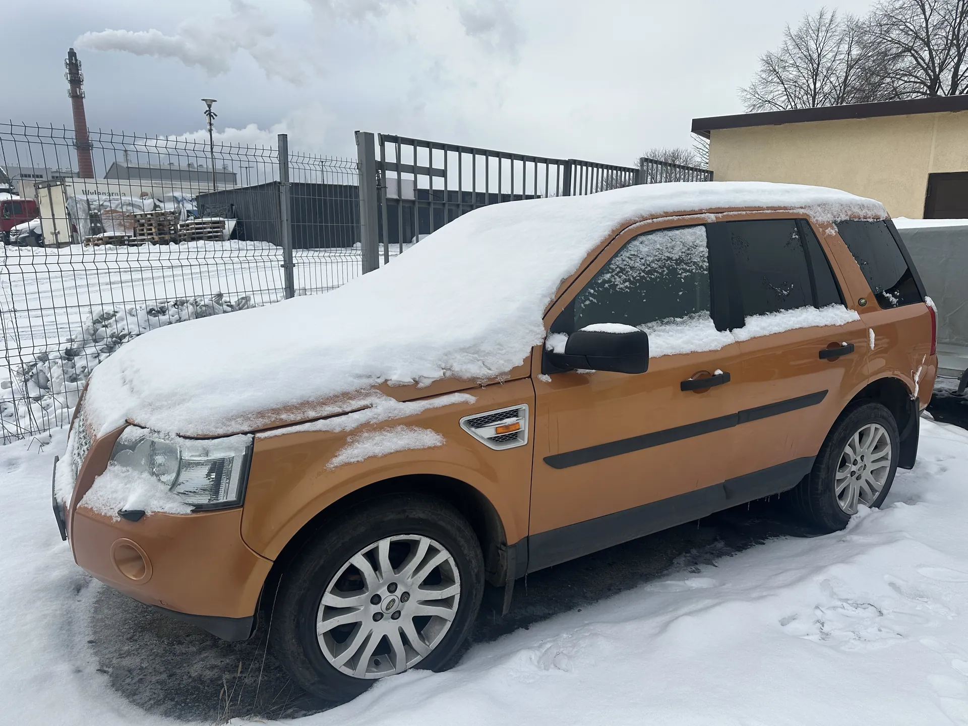 Brązowy SUV pokryty śniegiem zaparkowany na śnieżnej powierzchni. W tle metalowe ogrodzenie, przemysłowe budynki, komin z dymem, bezlistne drzewa oraz pochmurne niebo. Śnieg pokrywa dach i maskę pojazdu, a na ziemi widoczne są ślady. Koła samochodu mają srebrne felgi.
