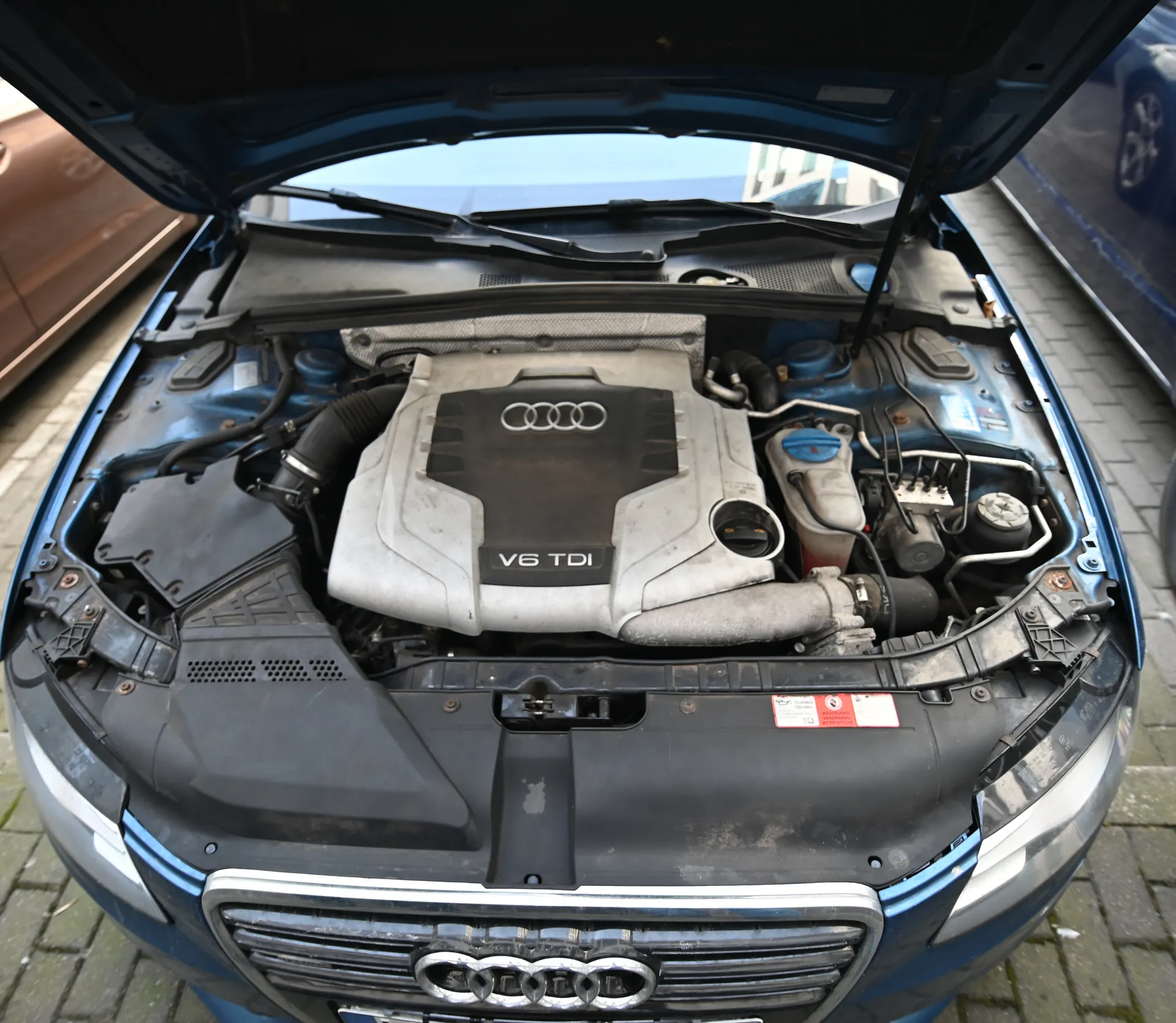 Samochód marki Audi z otwartą maską, ukazujący silnik z napisem V6 TDI i czterema pierścieniami logo Audi na pokrywie. Widoczne są elementy silnika, w tym zbiornik płynu chłodzącego z niebieską pokrywką oraz czerwono-biały etykietka ostrzegawcza. Przednia część pojazdu z charakterystyczną atrapą chłodnicy z logo Audi. W tle fragmenty innych samochodów i brukowana powierzchnia.