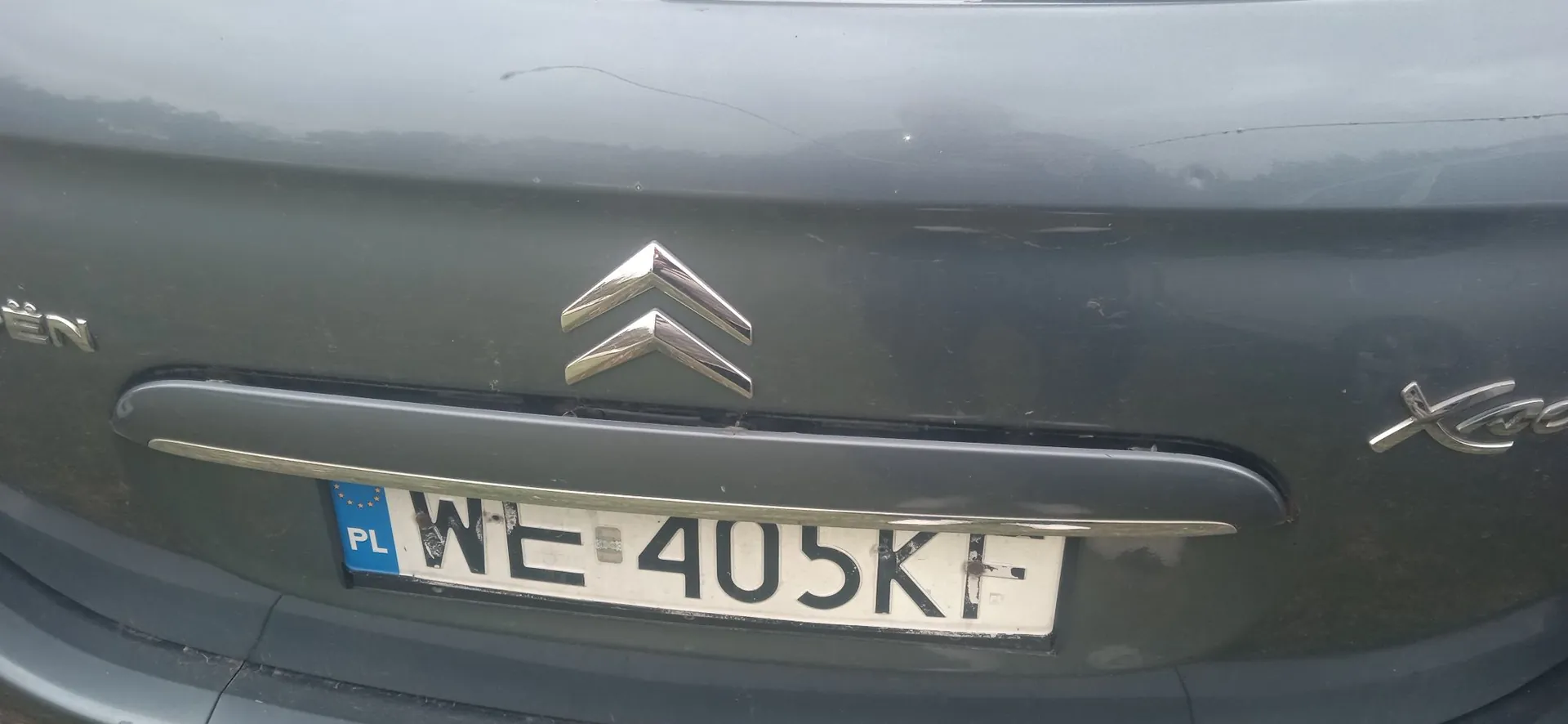 Zbliżenie na tylną część szarego samochodu marki Citroën. Na klapie bagażnika widoczne jest srebrne logo marki w kształcie dwóch odwróconych kątów. Poniżej znajduje się polska tablica rejestracyjna o numerze WE 405KF z niebieskim paskiem Unii Europejskiej i symbolem PL po lewej stronie.